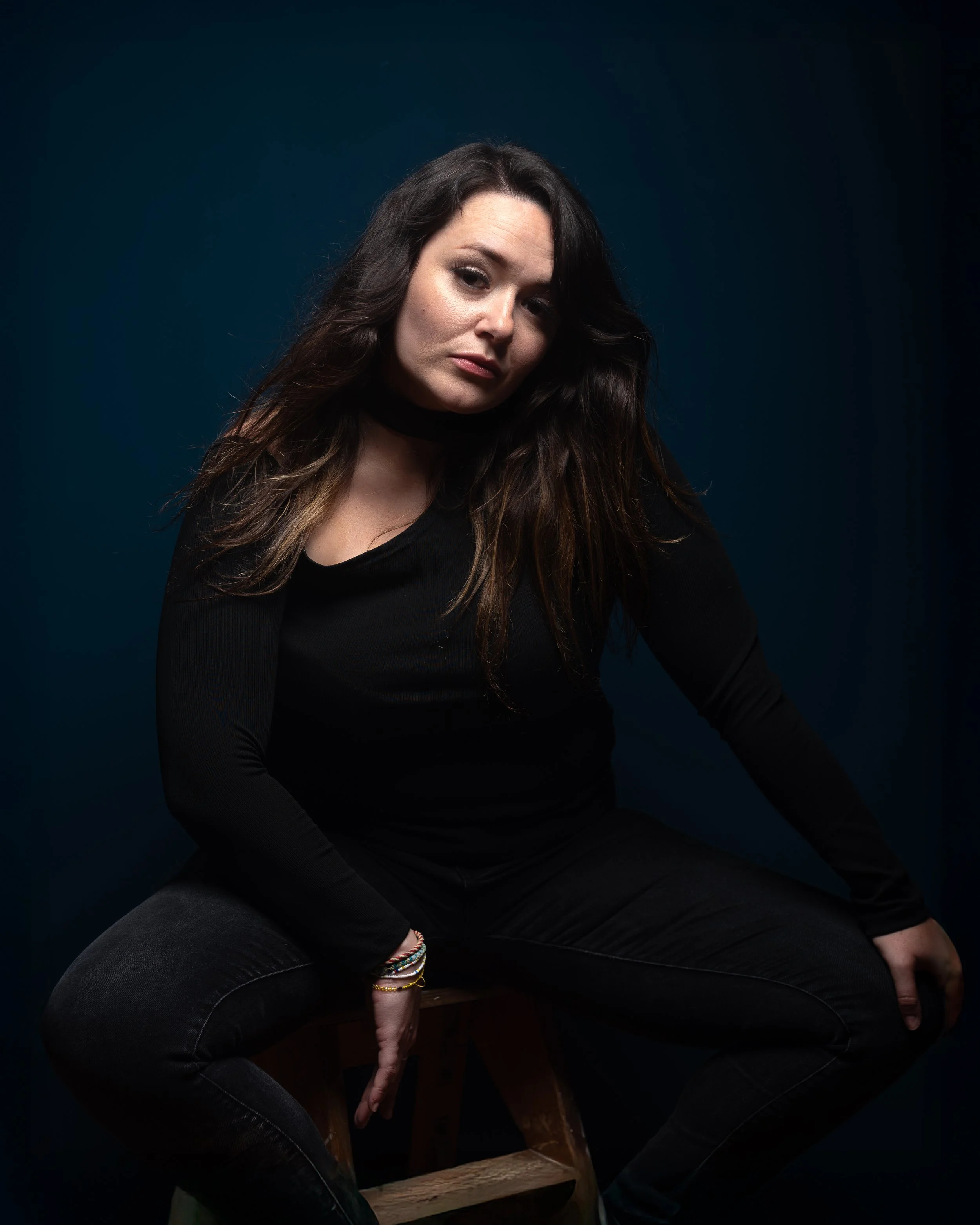 Une femme aux cheveux bruns foncés bouclés, portant un haut noir, assise sur une chaise en bois, contre un fond sombre. Elle regarde légèrement vers la caméra avec une expression neutre.