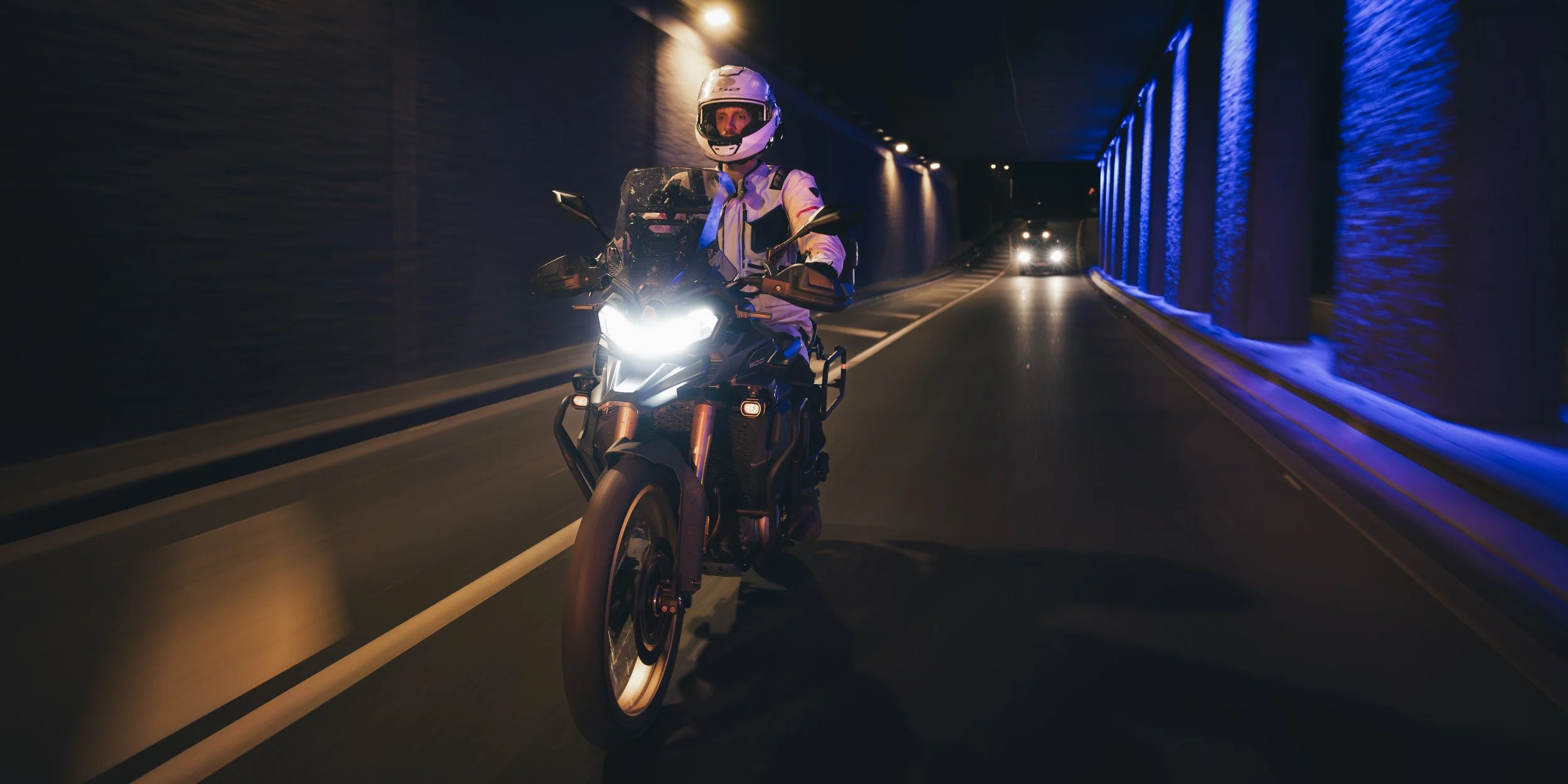 Motard portant un casque et une tenue de moto, roulant dans un tunnel la nuit, avec d'autres véhicules en arrière-plan.