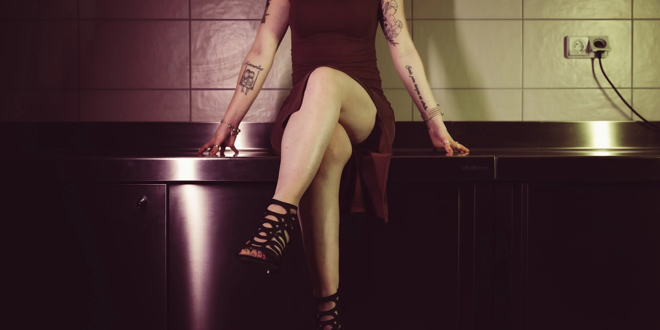Une femme assise sur une surface en métal dans une pièce, portant une robe marron, des chaussures à talons ouvertes. Ses bras sont tatoués et elle porte plusieurs bijoux.