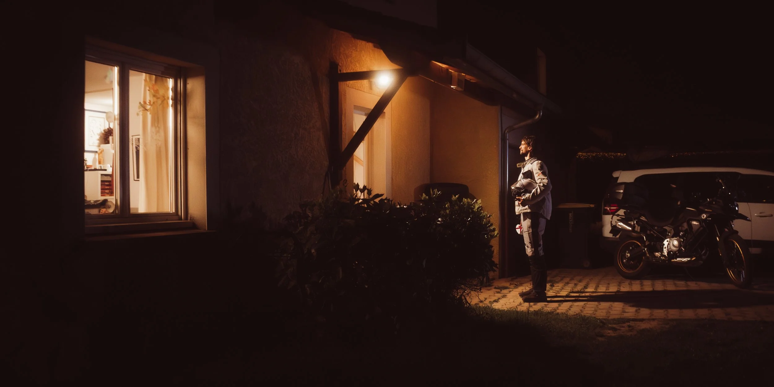 Un homme se tient devant une maison la nuit, éclairé par une lampe, avec des voitures et une moto garées à proximité.