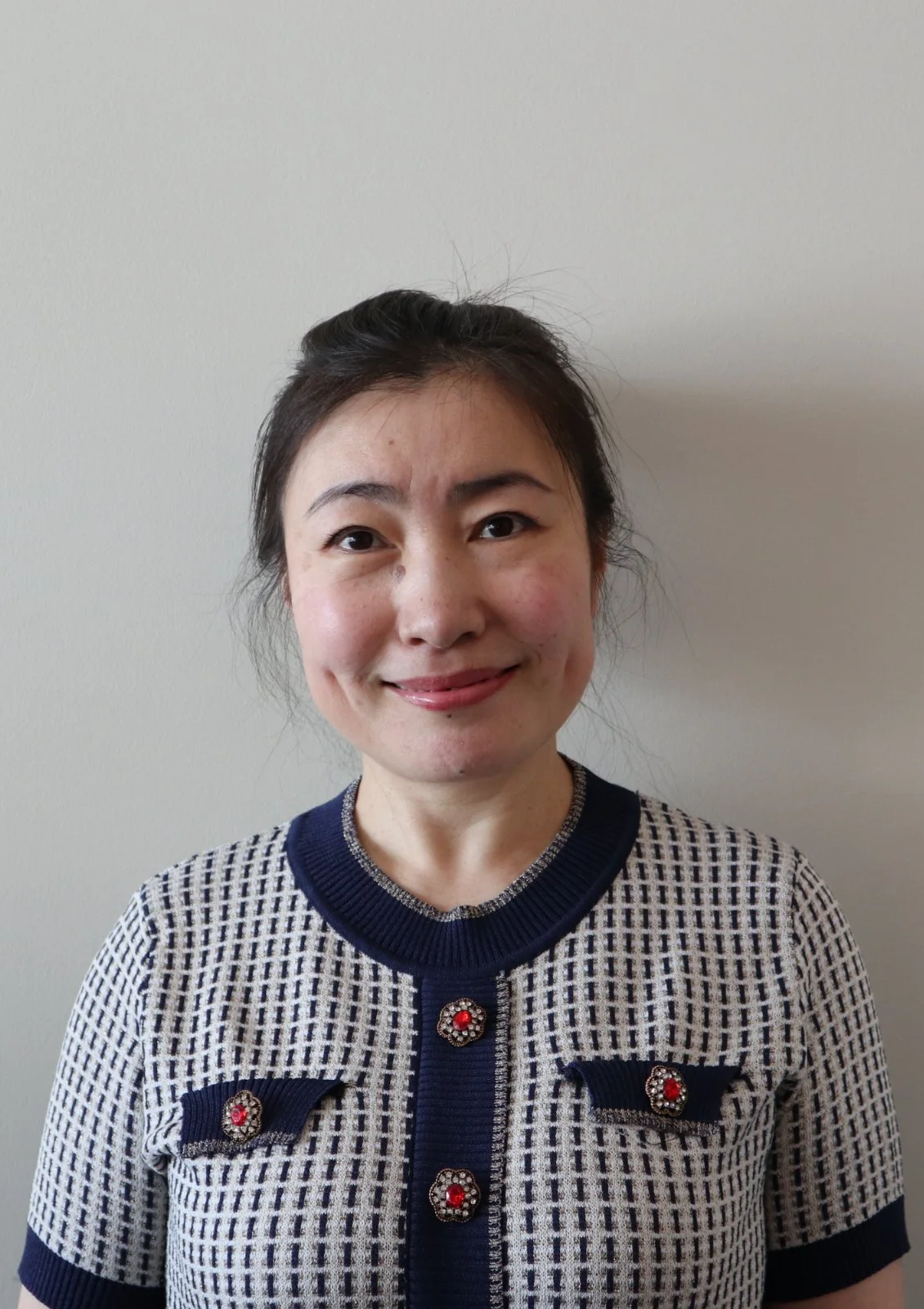 A portrait of a smiling Asian woman with dark hair in a ponytail, wearing a patterned navy and white sweater with decorative buttons and bows.
