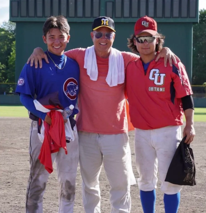 Three people standing together on a baseball field, smiling with arms around each other. Two are wearing baseball uniforms, and the middle person is dressed casually with sunglasses and a hat.