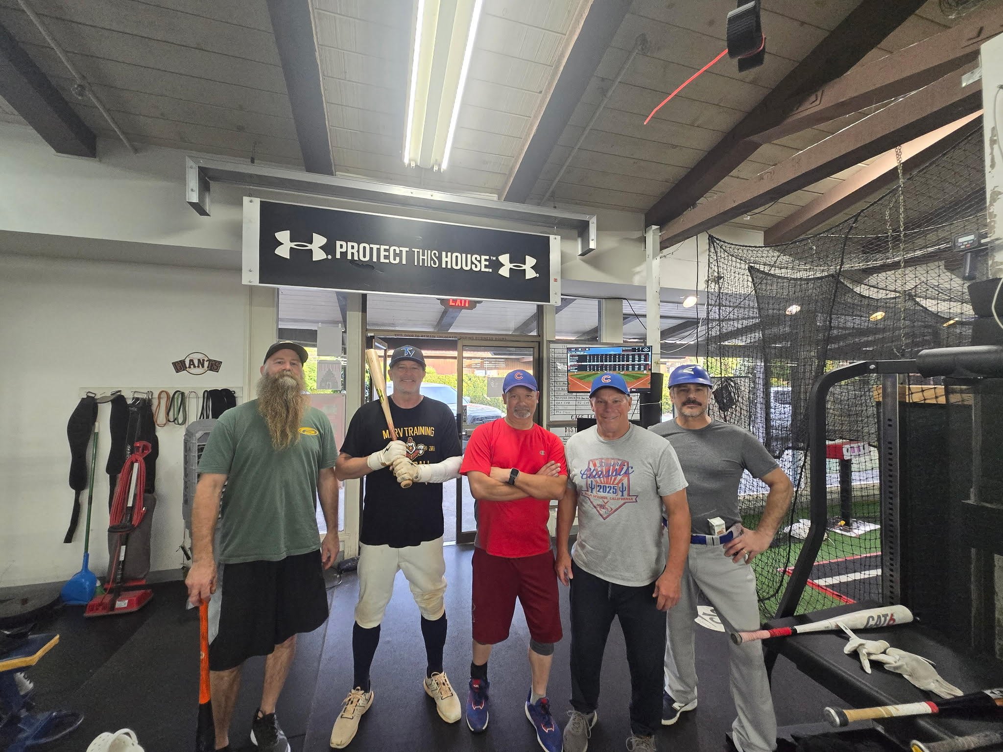 A group of five men standing together inside a gym. They are wearing athletic clothes and caps, and are posing for the photo. Behind them, there is gym equipment, a netted area, and a sign that reads 'PROTECT THIS HOUSE' with the Under Armour logo.