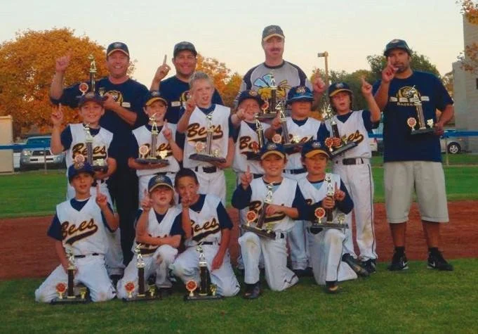 Youth baseball team with coaches celebrating, holding trophies, in uniforms, on a baseball field during fall, with autumn trees in the background.