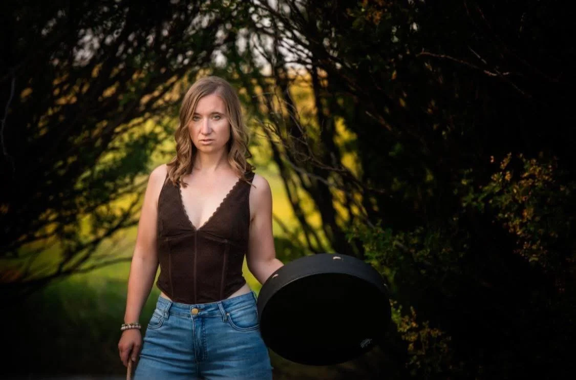 A young woman with blonde hair standing outdoors in a wooded area during sunset, holding a black circular object, wearing a dark sleeveless top and blue jeans.