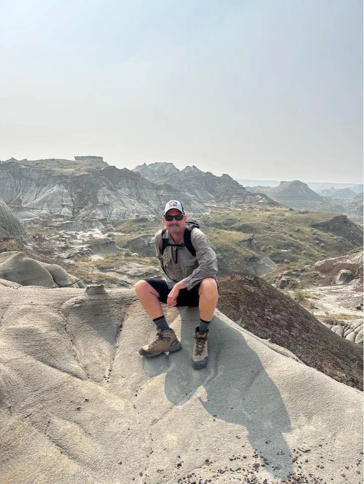 A man sitting on a rock formation in a desert landscape with rugged hills and rock formations in the background.