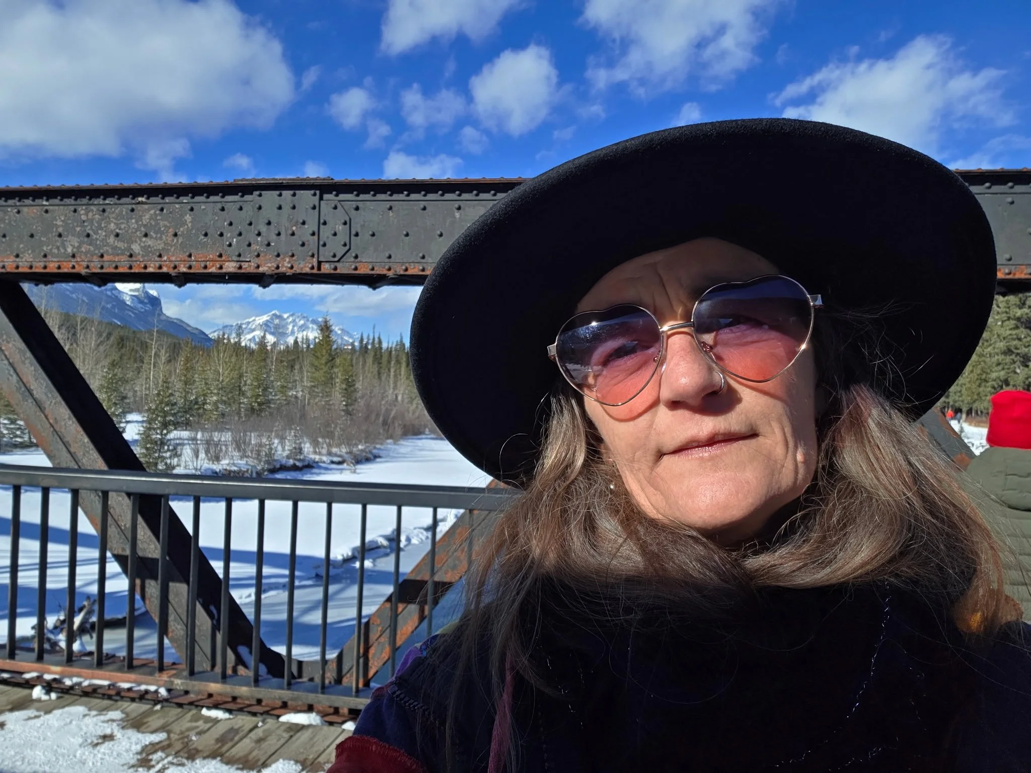 A woman with long hair wearing a wide-brimmed black hat and sunglasses, standing outdoors on a snow-covered bridge with a mountain range in the background under a partly cloudy sky.