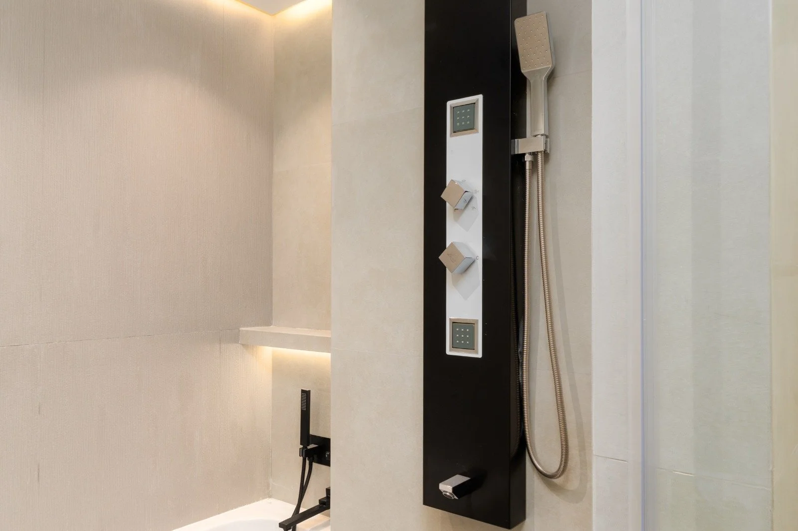 Modern shower panel with digital display and multiple controls, attached to a wall in a contemporary bathroom with beige tiled walls.