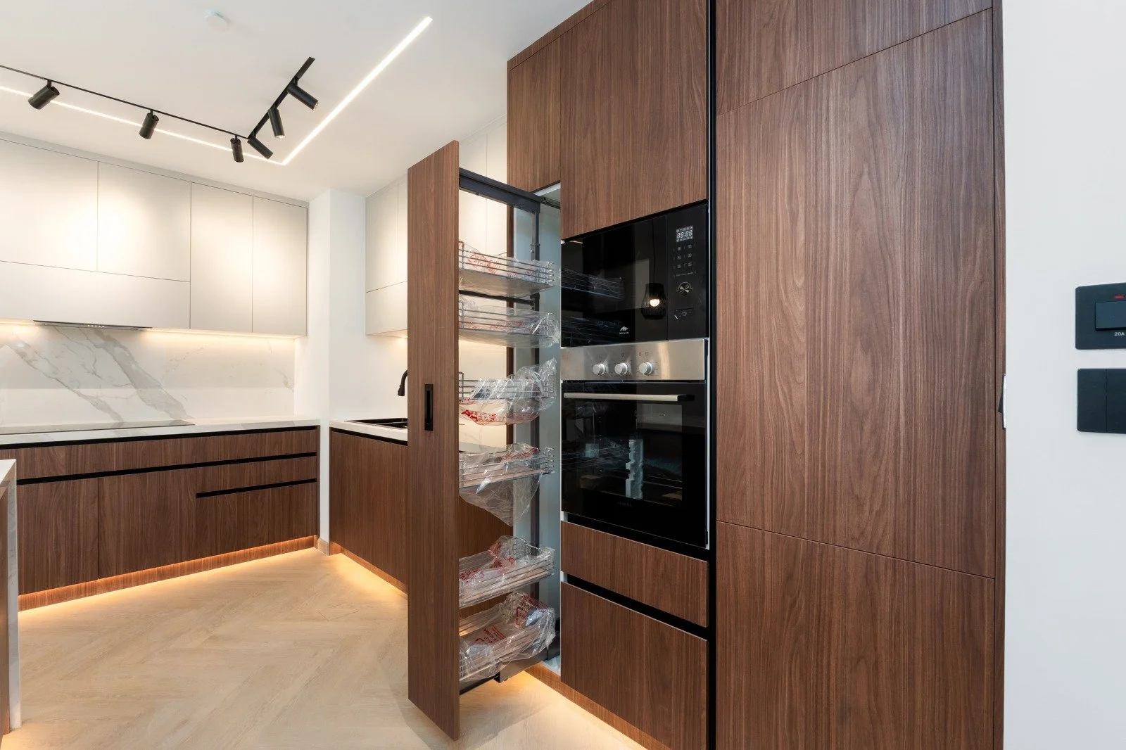 Modern kitchen with white cabinets, marble backsplash, black track lighting, and tall wooden pantry. Pull-out shelves in pantry contain food items, and wall-mounted oven and microwave are integrated into wooden cabinetry.