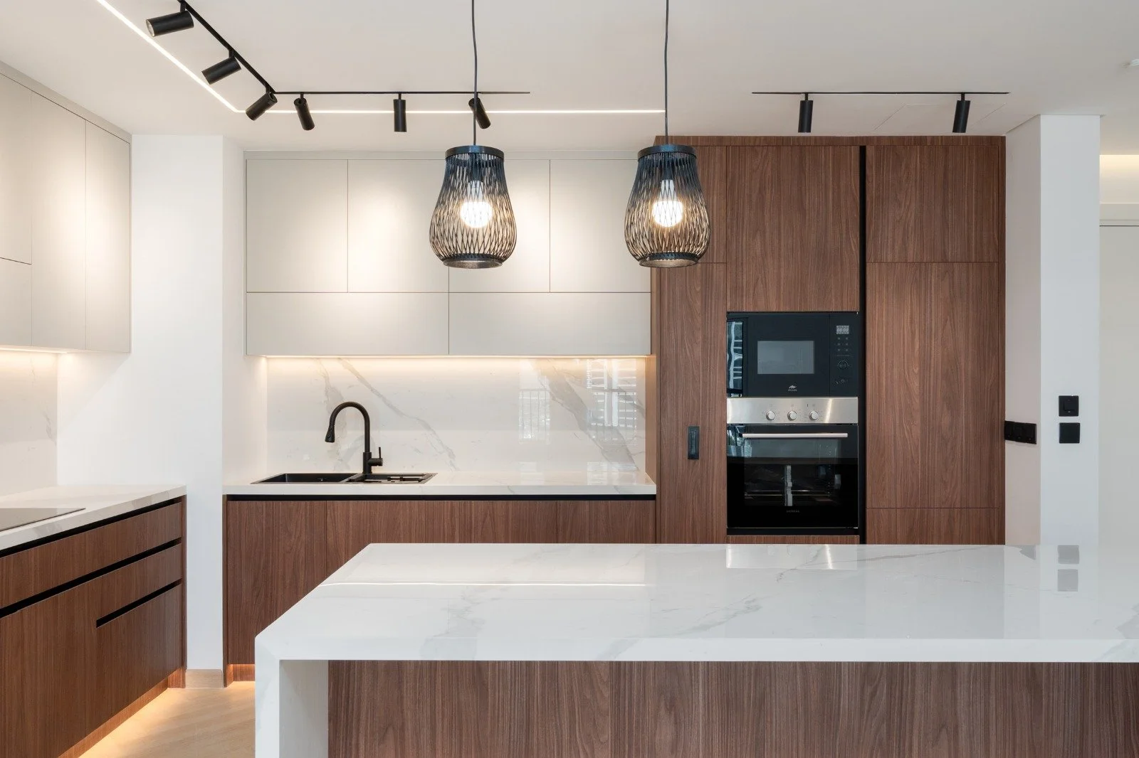 Modern kitchen with white marble countertops, wood cabinetry, black sink and faucet, and black appliances, illuminated by pendant lights and track lighting.