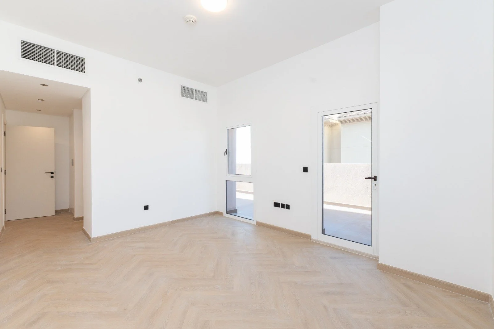Empty room with light-colored wooden flooring and white walls, featuring a door leading outside, a window, and another door leading inside, with vents and a smoke detector on the ceiling.