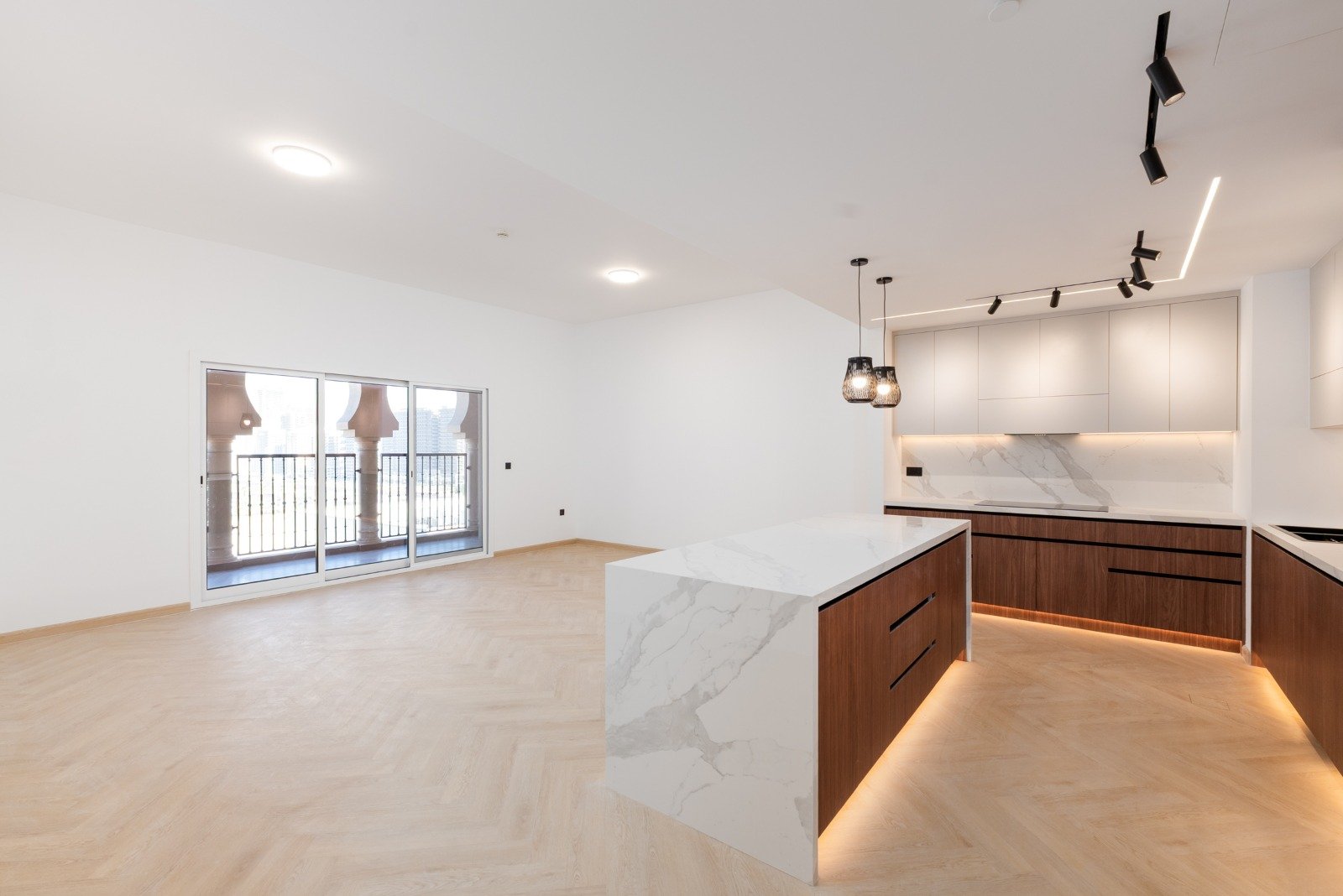 Modern kitchen with wooden cabinets, white marble countertops, a kitchen island, pendant lighting, and track lighting, opening to a bright living space with a glass sliding door and balcony.