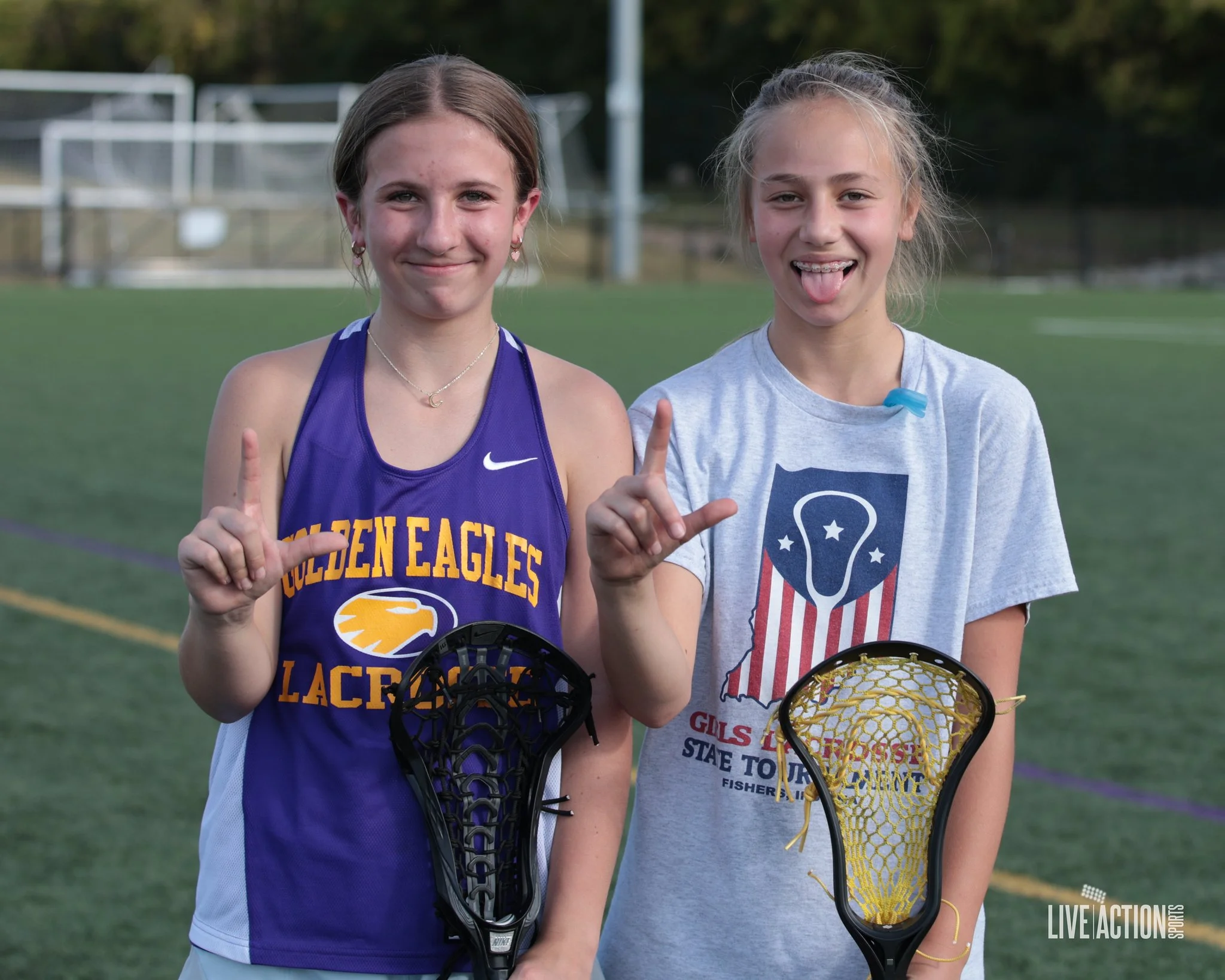 Junior Golden Eagles girls standing on a sports field, holding lacrosse sticks, smiling and making 'L' signs with their fingers.
