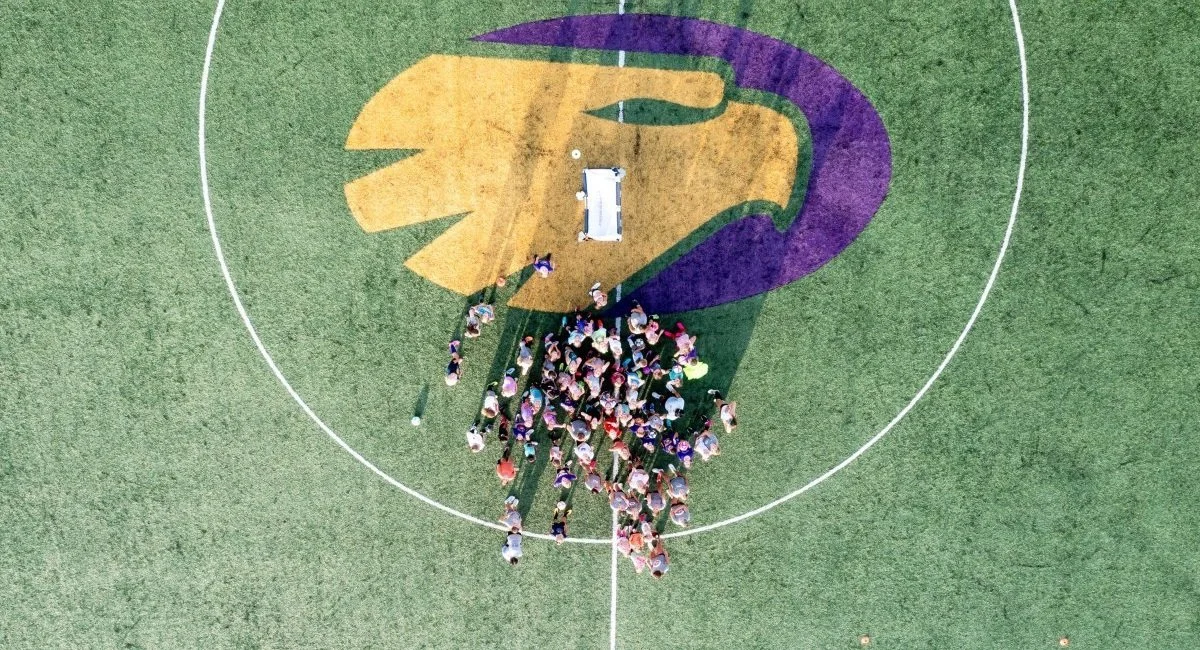 Junior Golden Eagles Youth Sports. A bird's-eye view of a group of children gathered on a sports field, standing on a large colorful logo painted on the ground that features a purple and yellow eagle head design.