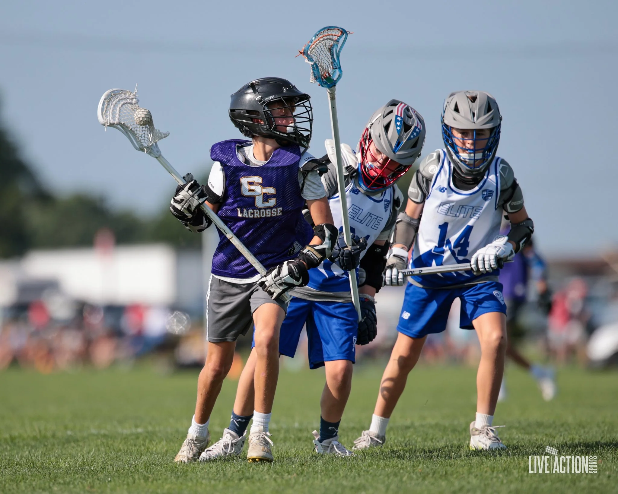 Junior Golden Eagles boys playing lacrosse outdoors on a grassy field, wearing helmets and athletic gear, with a crowd in the background.