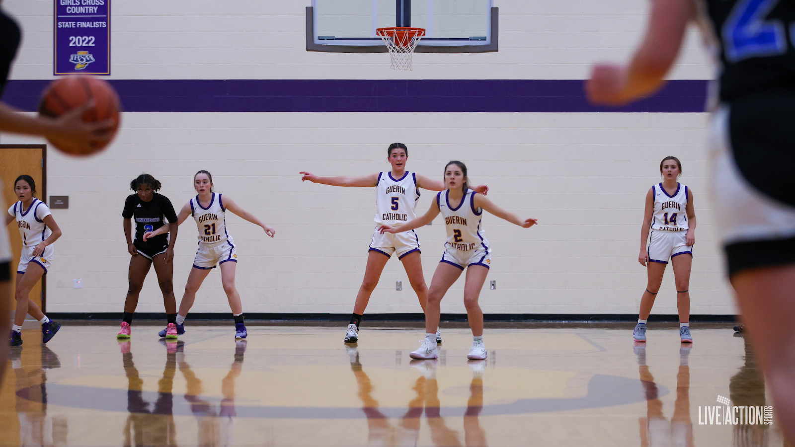 Junior Golden Eagles Girls' basketball team in white and purple uniforms on the court during a game, with a basketball hoop in the background.