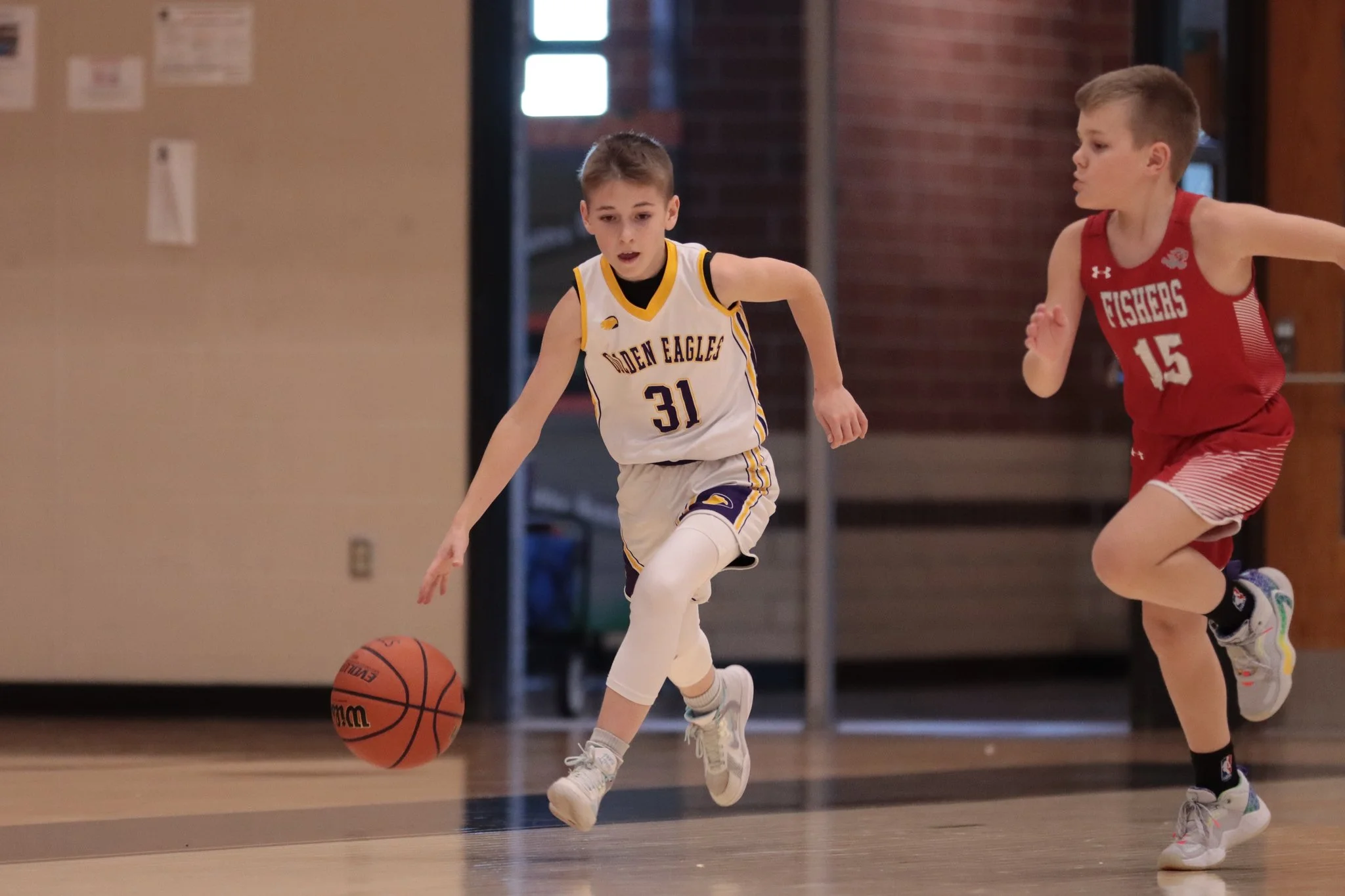 Junior Golden Eagles boys playing basketball indoors. One boy, wearing a white and yellow jersey with the number 31, is dribbling the ball. The other boy, wearing a red jersey with the number 15, is running alongside him.