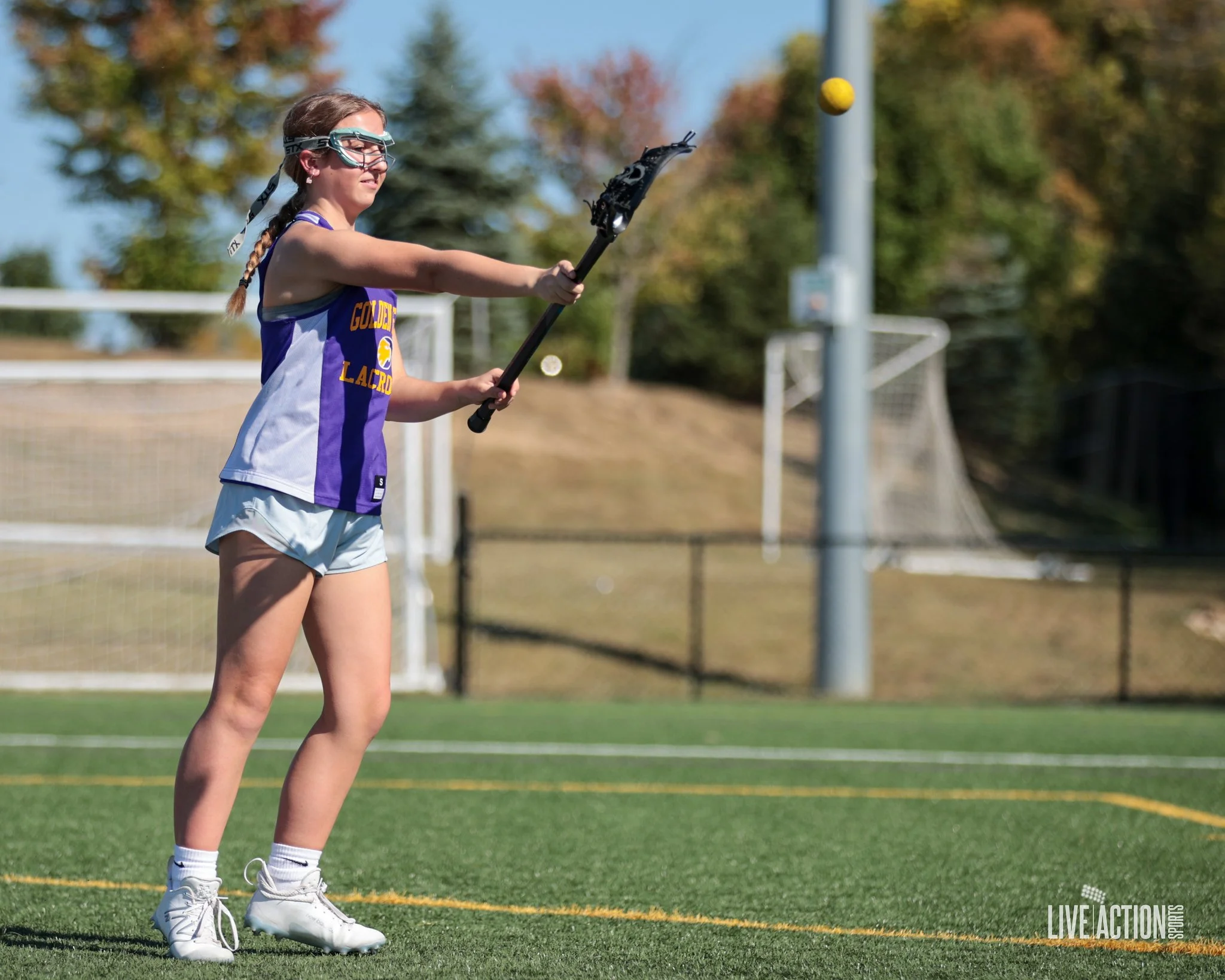 A female lacrosse player in a purple and white jersey and light shorts is on a grassy field, preparing to catch or pass a yellow ball with her lacrosse stick. She is wearing protective goggles and is focused on the game, with trees and sports goals in the background.
