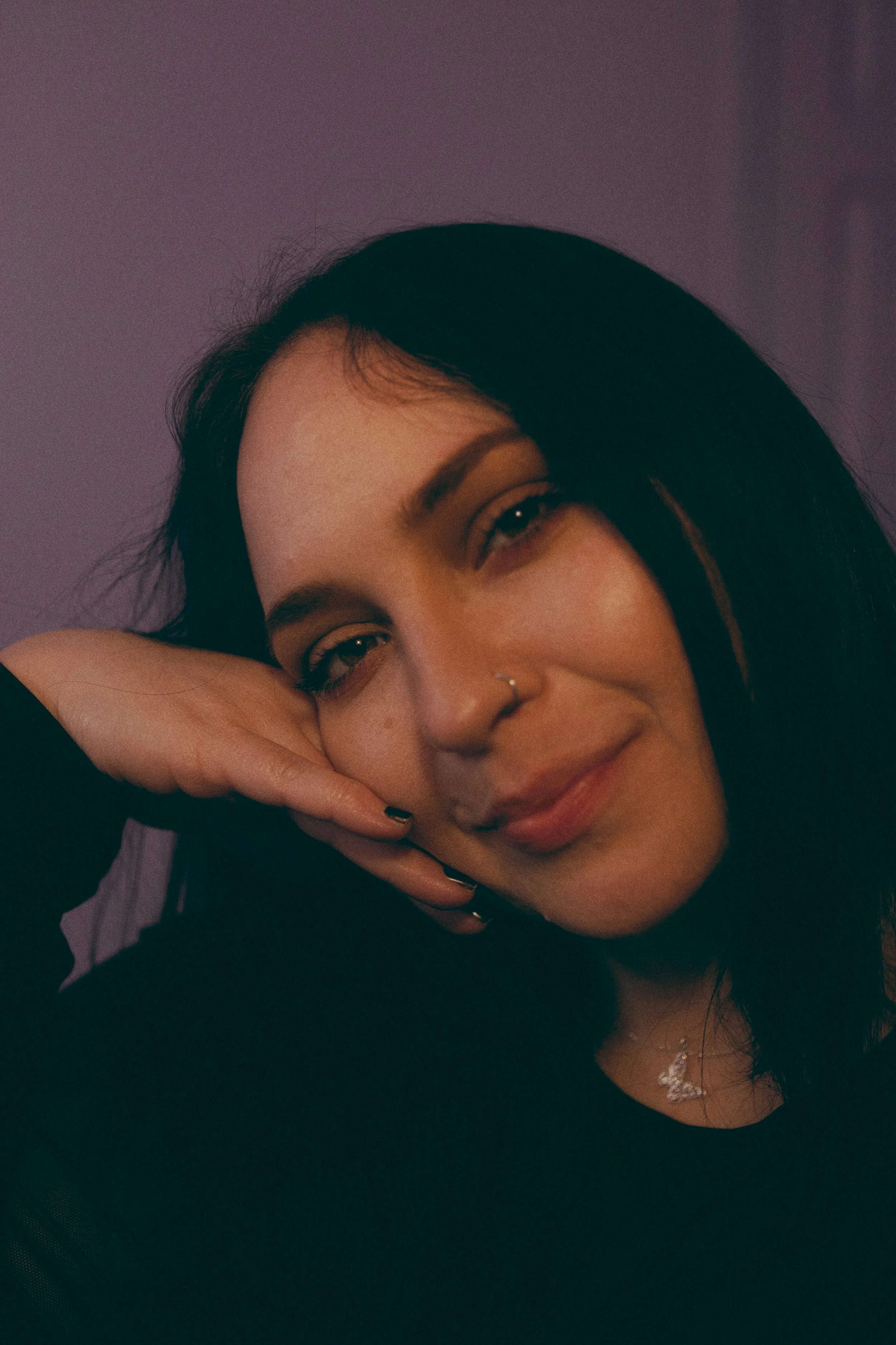Close-up of a young woman with black hair, nose ring, and a necklace, smiling gently with one hand resting on her face against a purple background.