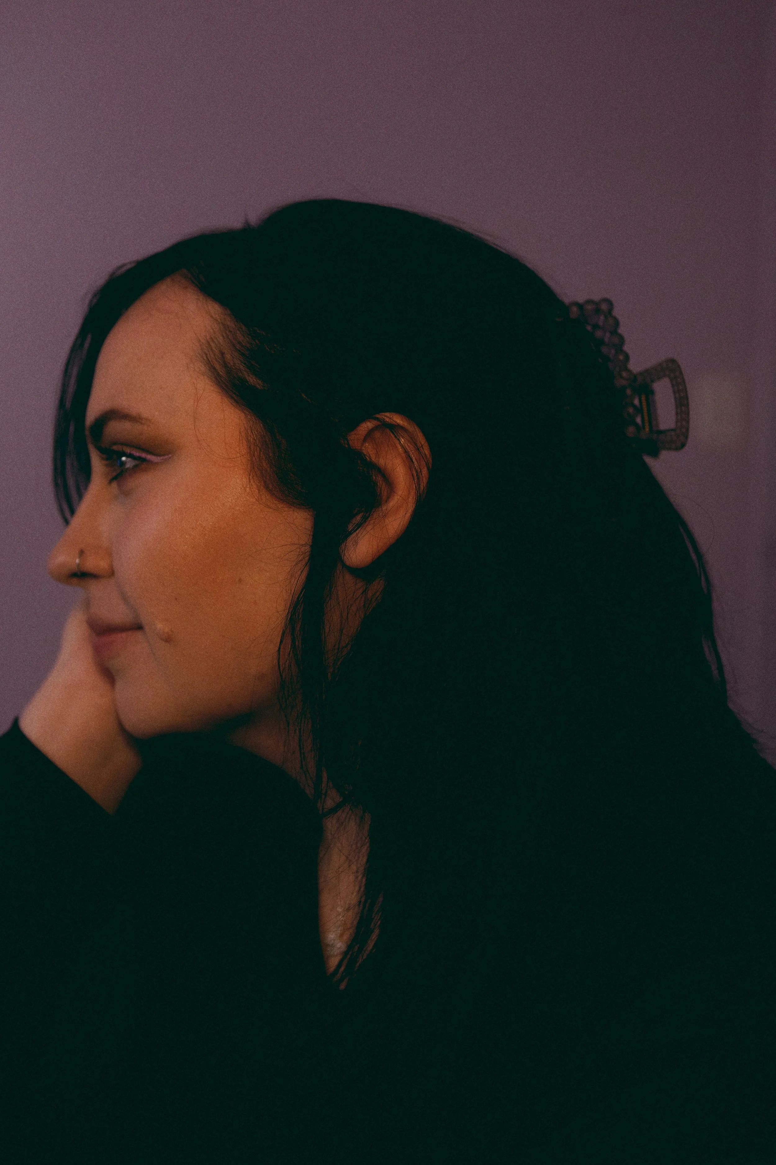 A woman with dark hair, wearing makeup, a nose ring, and a hair clip, resting her face on her hand, against a purple background.