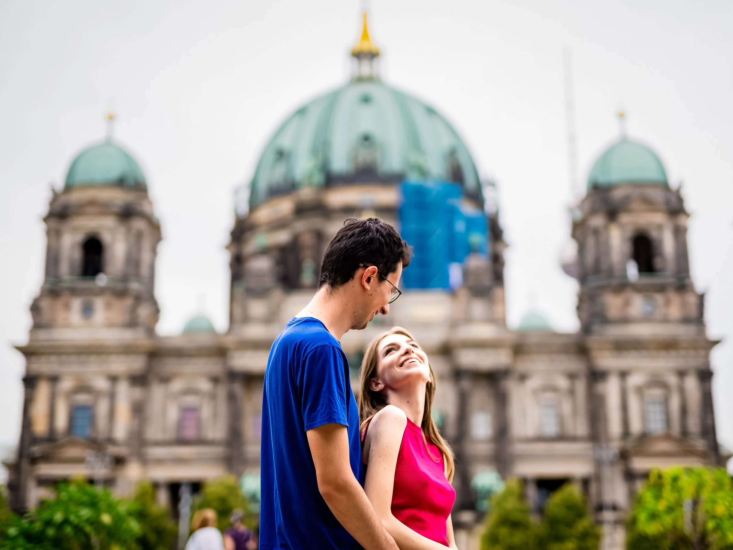 A close-up of a young couple with their faces touching, looking at the camera. The man has light brown hair, blue eyes, and a slight beard, while the woman has red hair, fair skin, and freckles.