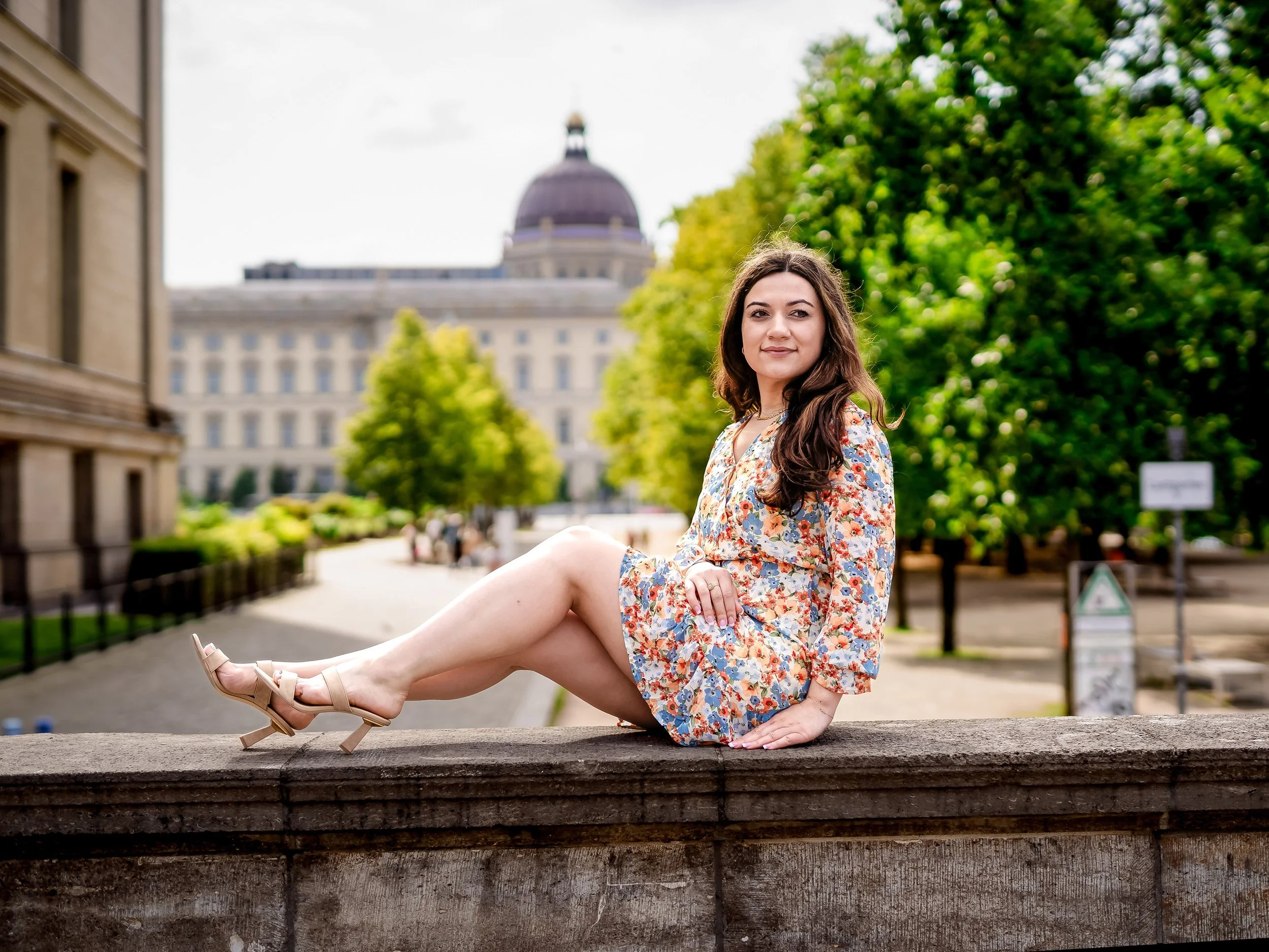 A young woman in a wedding dress holding a bouquet of pink and white roses, standing in a floral decorated venue with blurred background of flowers and lights.