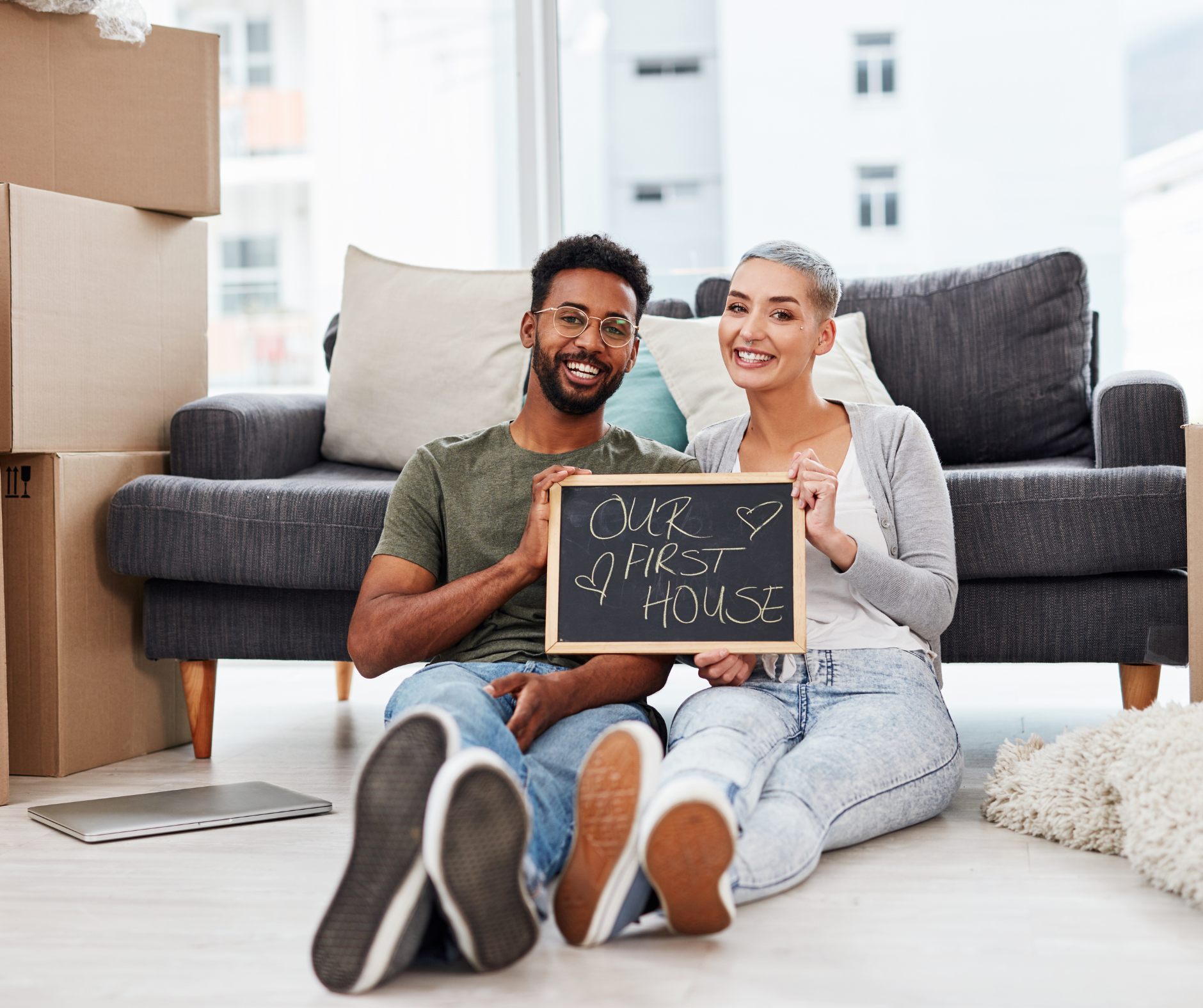 Happy couple sitting on the floor of new apartment with moving boxes, holding a chalkboard sign that says 'Our First House' with hearts.