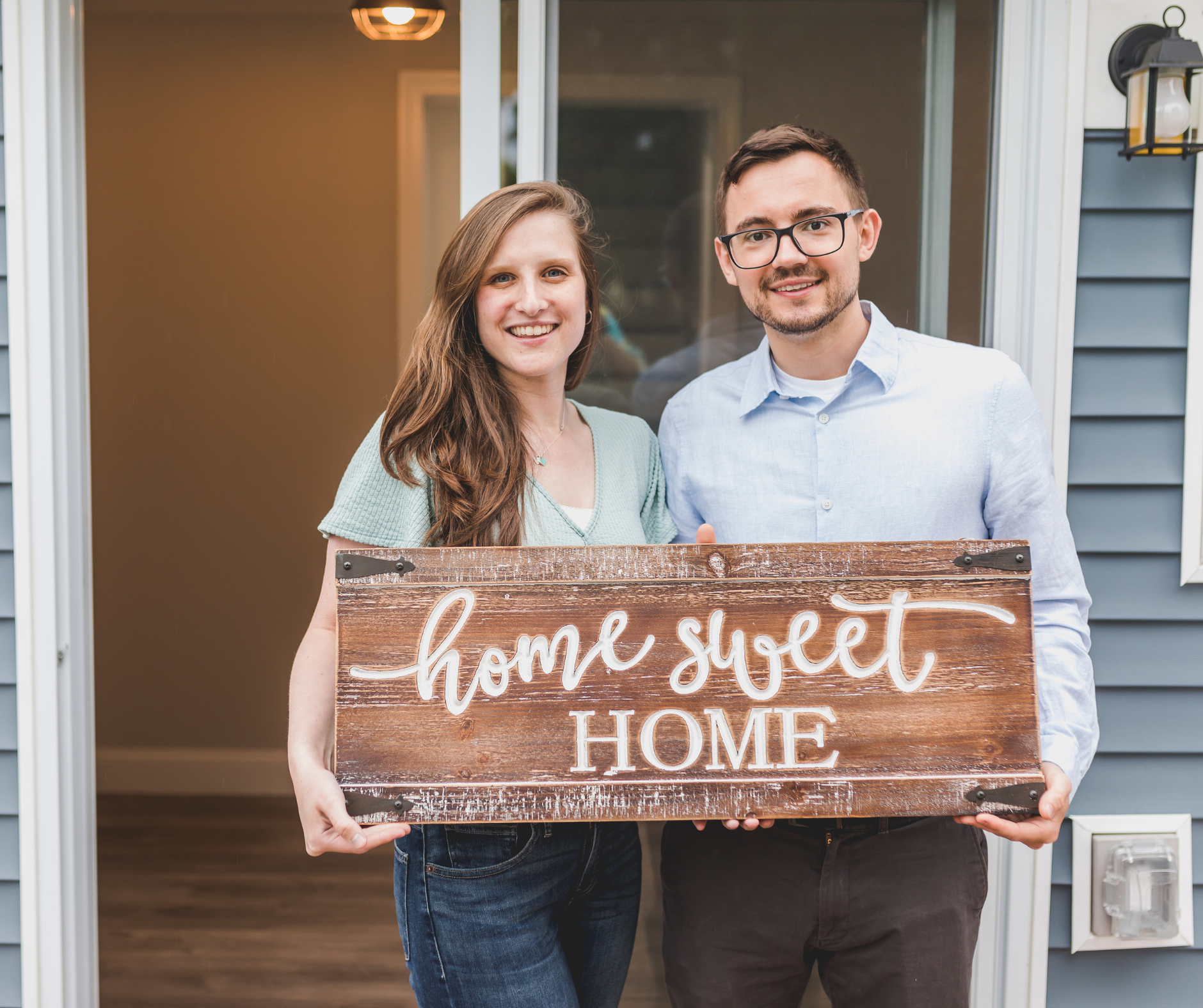 A smiling couple standing at the entrance of a house, holding a wooden sign that says "home sweet home."