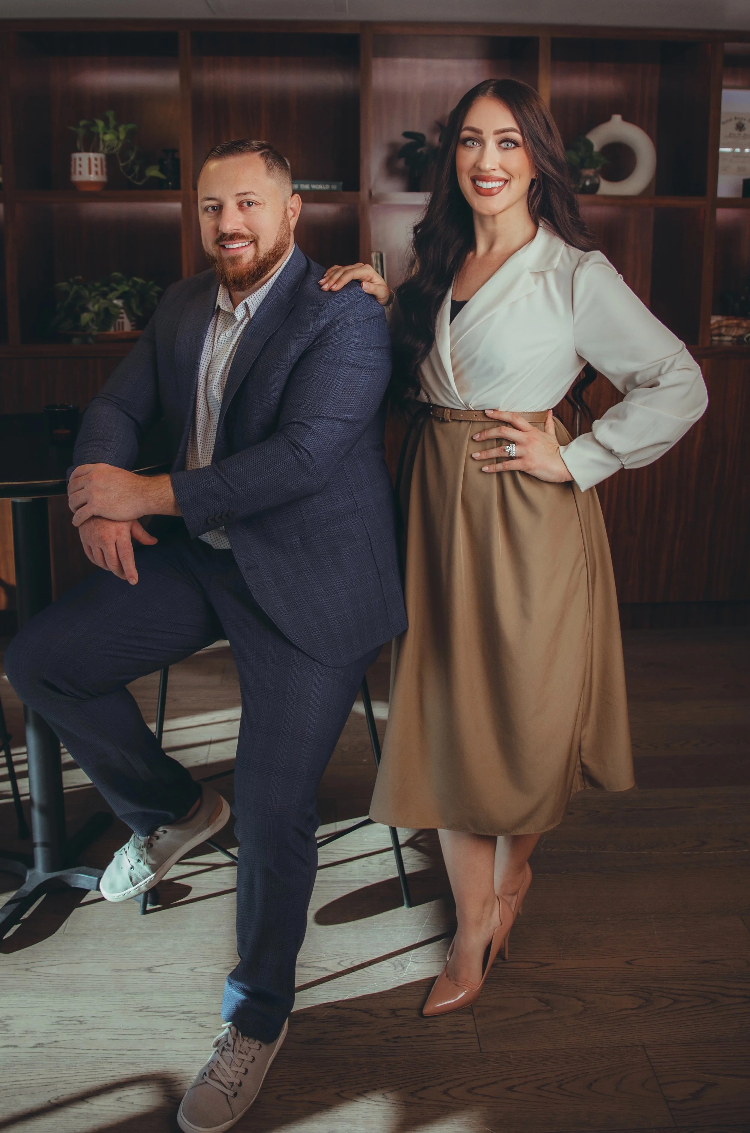 A man in a blue suit sitting on a stool and a woman in a beige skirt and white blouse standing beside him with her hand on his shoulder, in a room with dark wooden shelves and decorative items.