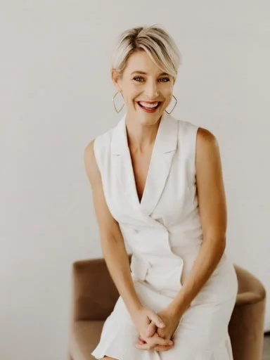 A smiling woman with short blonde hair, wearing a sleeveless white vest, sitting on a brown chair against a plain light background.