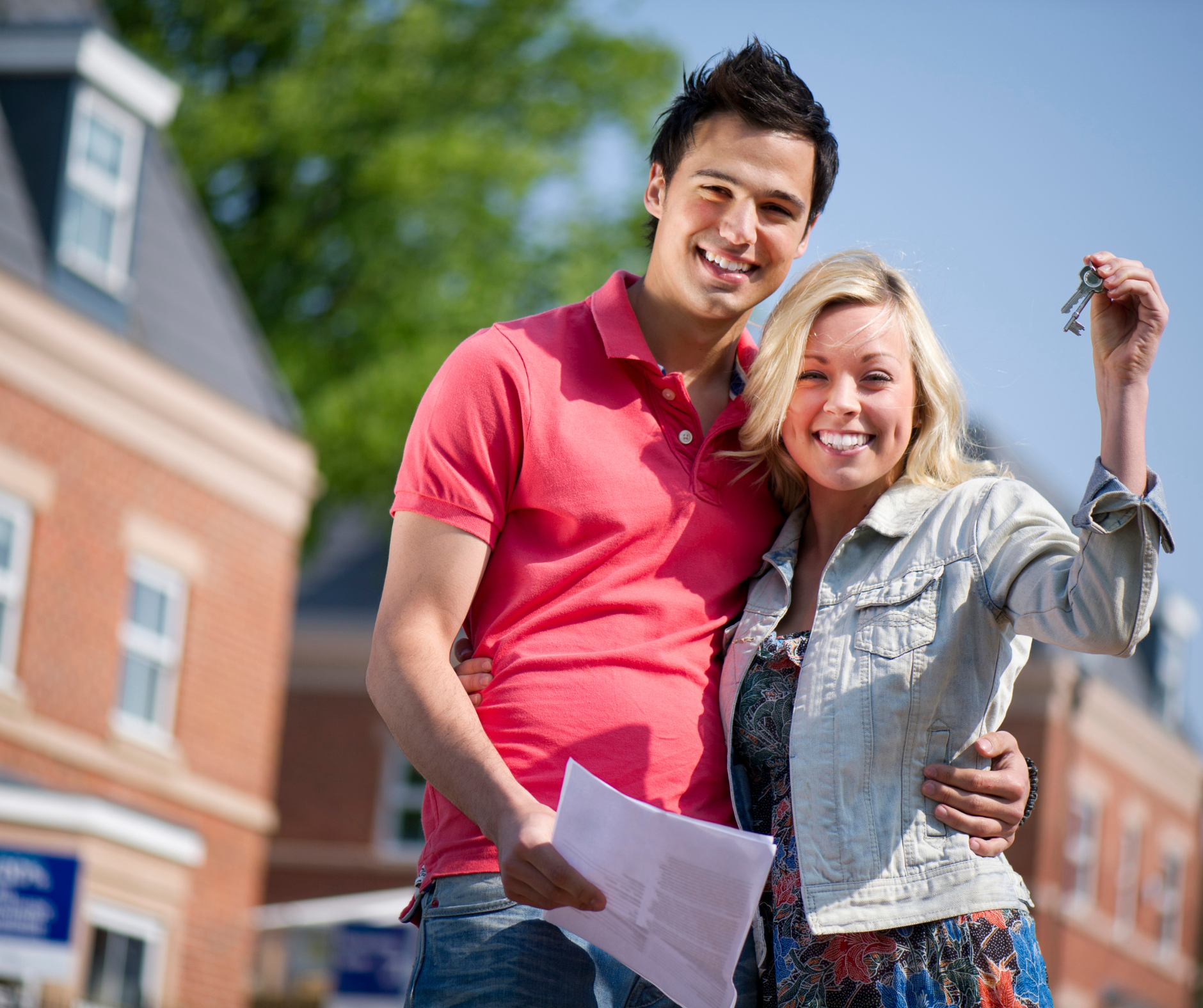 A happy young couple standing outdoors, with the woman holding up keys and a document, in front of houses, on a sunny day.