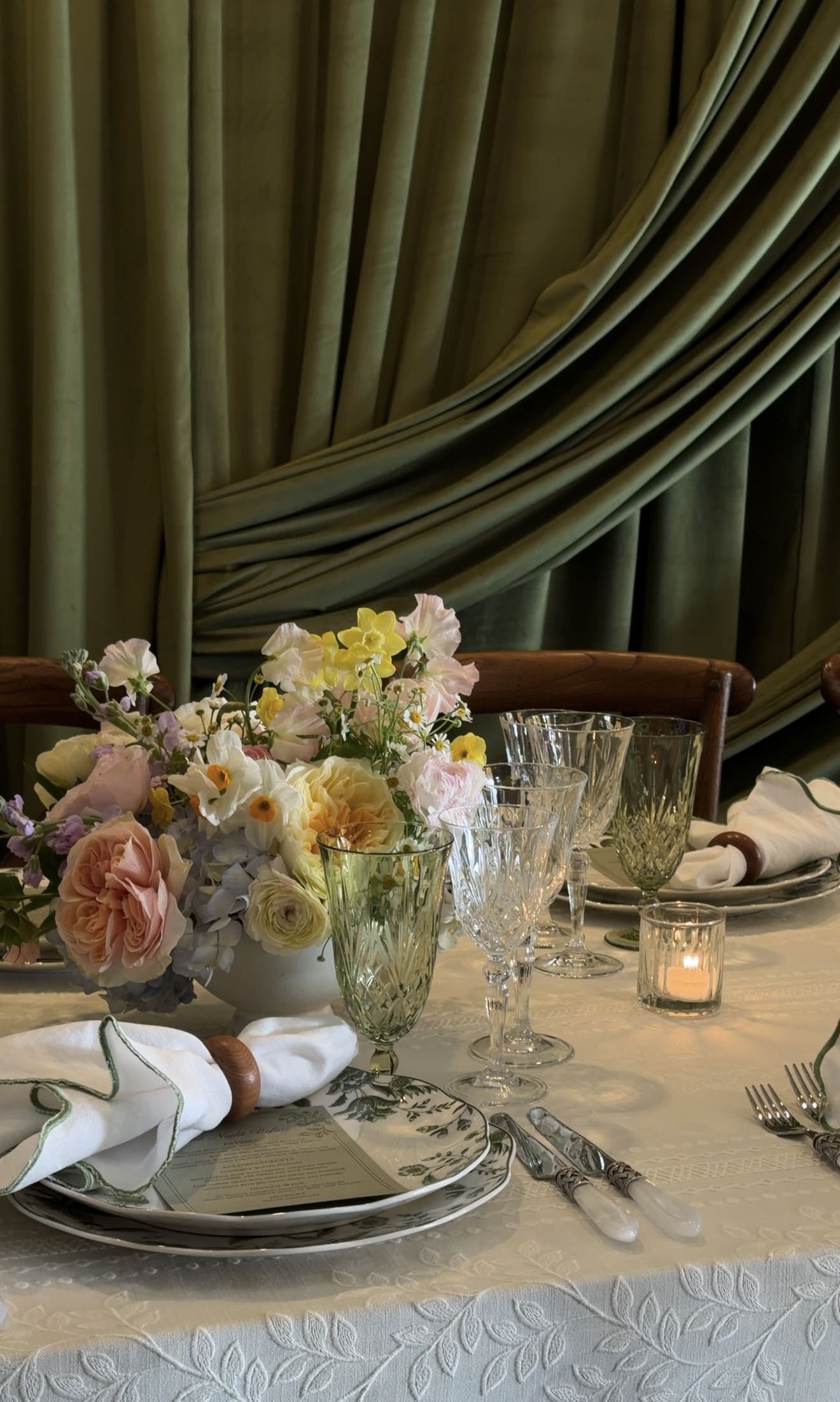 Elegant dining table set with floral centerpiece, crystal glasses, white plates, silverware, and white napkins on a tablecloth. Green velvet curtains in the background.