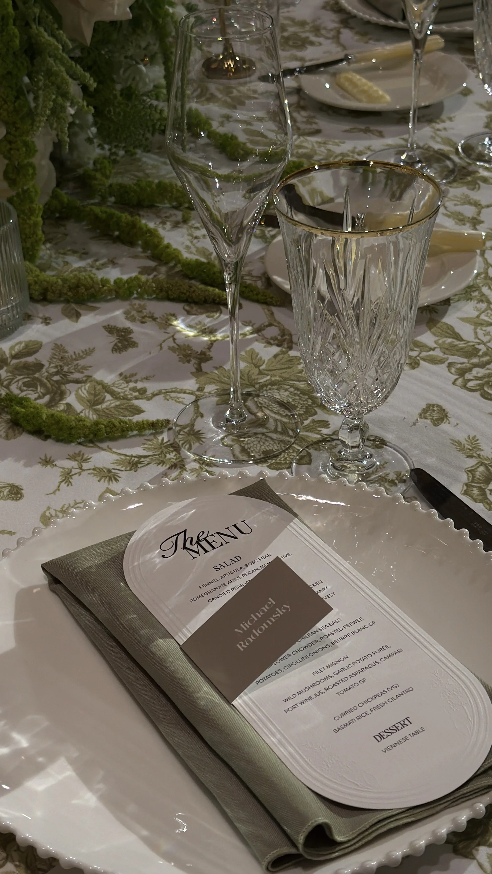 A formal dining table set with glassware, a menu, a napkin, and decorative green garland on a green and gold patterned tablecloth.