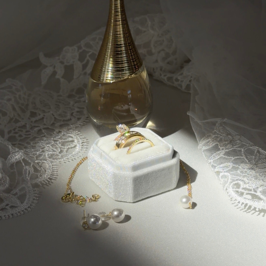 A jewelry display with rings, earrings, and a gold necklace, set on a white surface with lace fabric and a decorative bottle in the background.