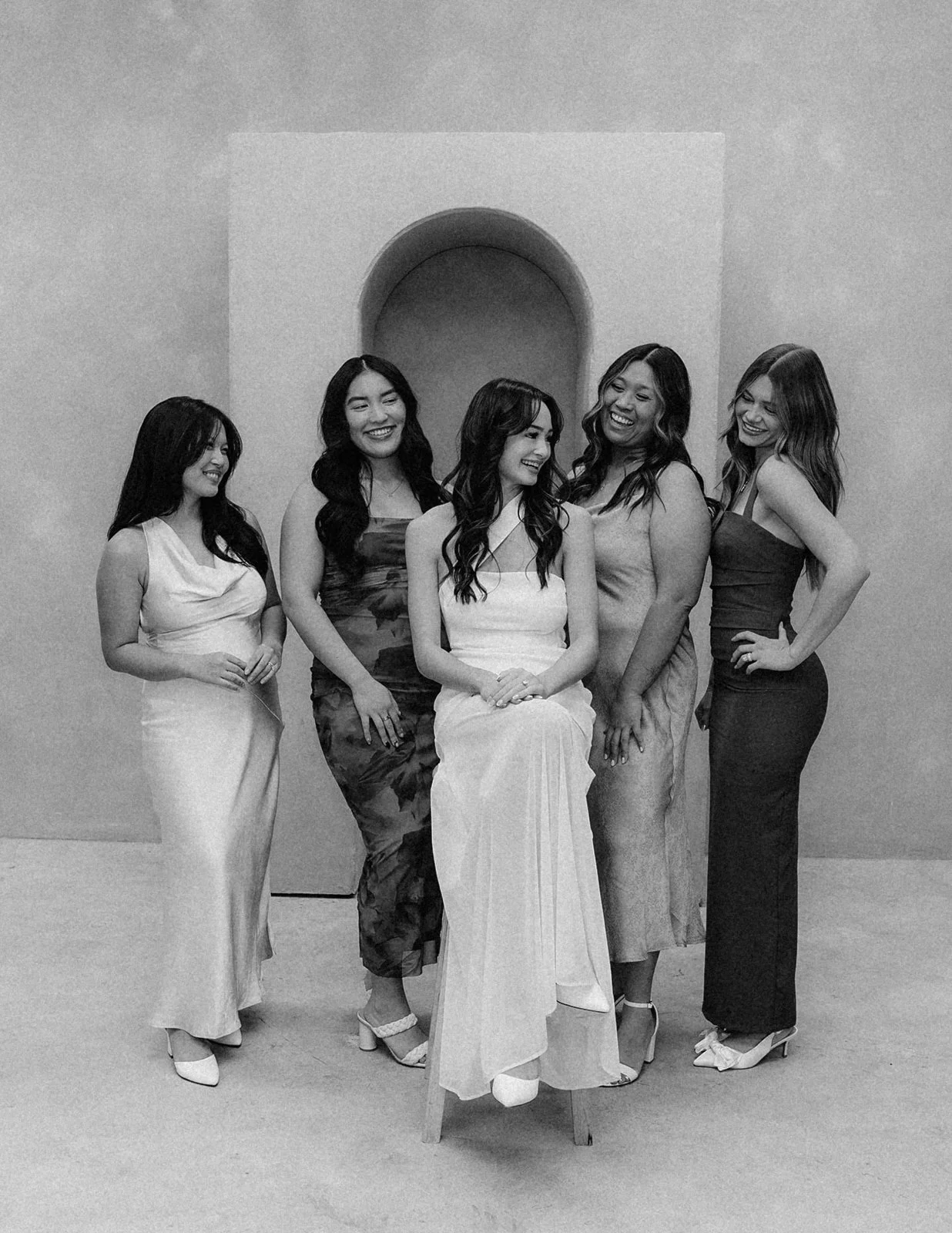 A group of five women in evening dresses posing together in a gallery or museum with a plain wall and a small arch in the background. One woman is sitting on a chair, while the others stand around her, all smiling and engaging with each other.