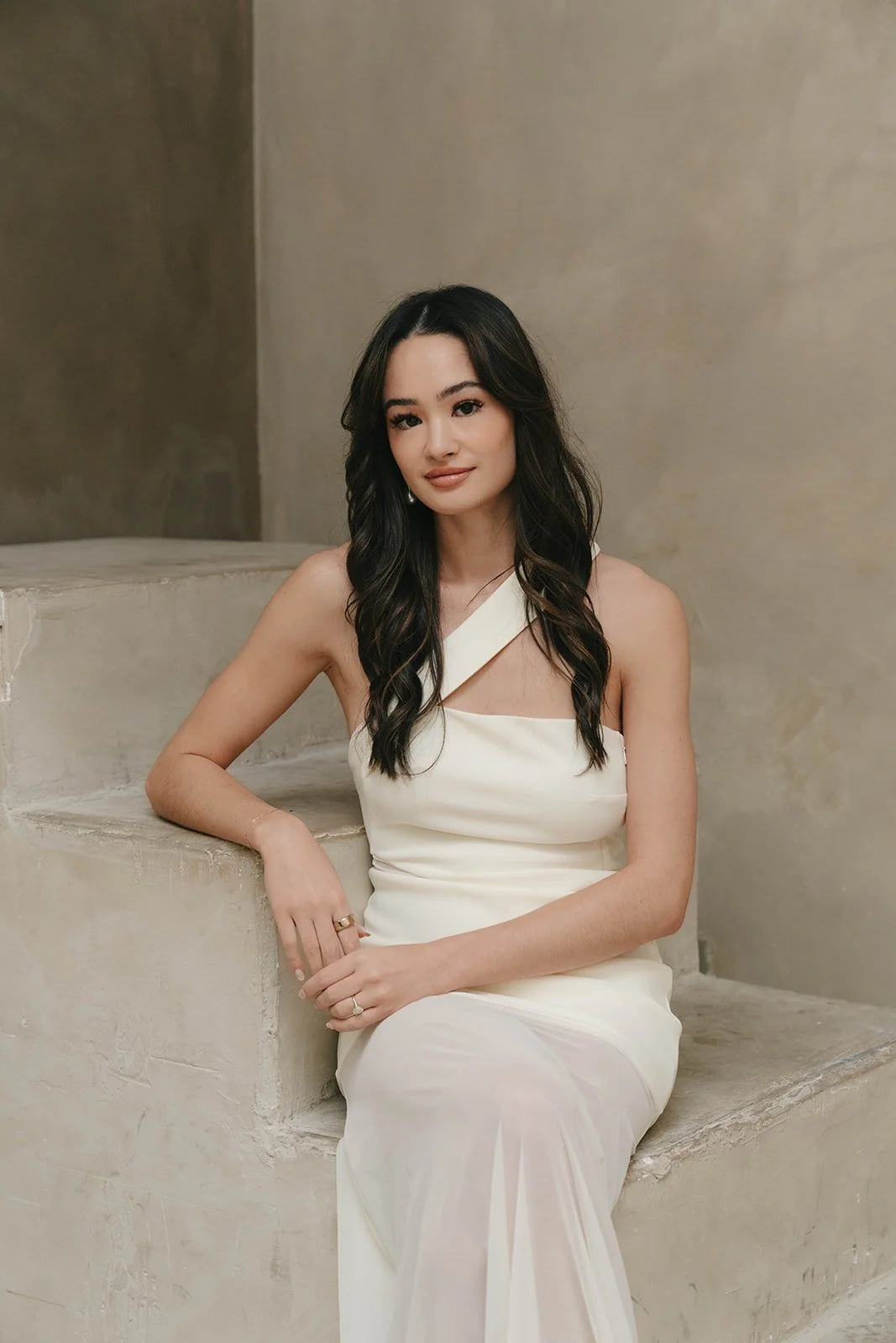 A young woman with long dark hair in loose waves, wearing a white one-shoulder dress, sitting on concrete steps in an indoor setting with beige walls.