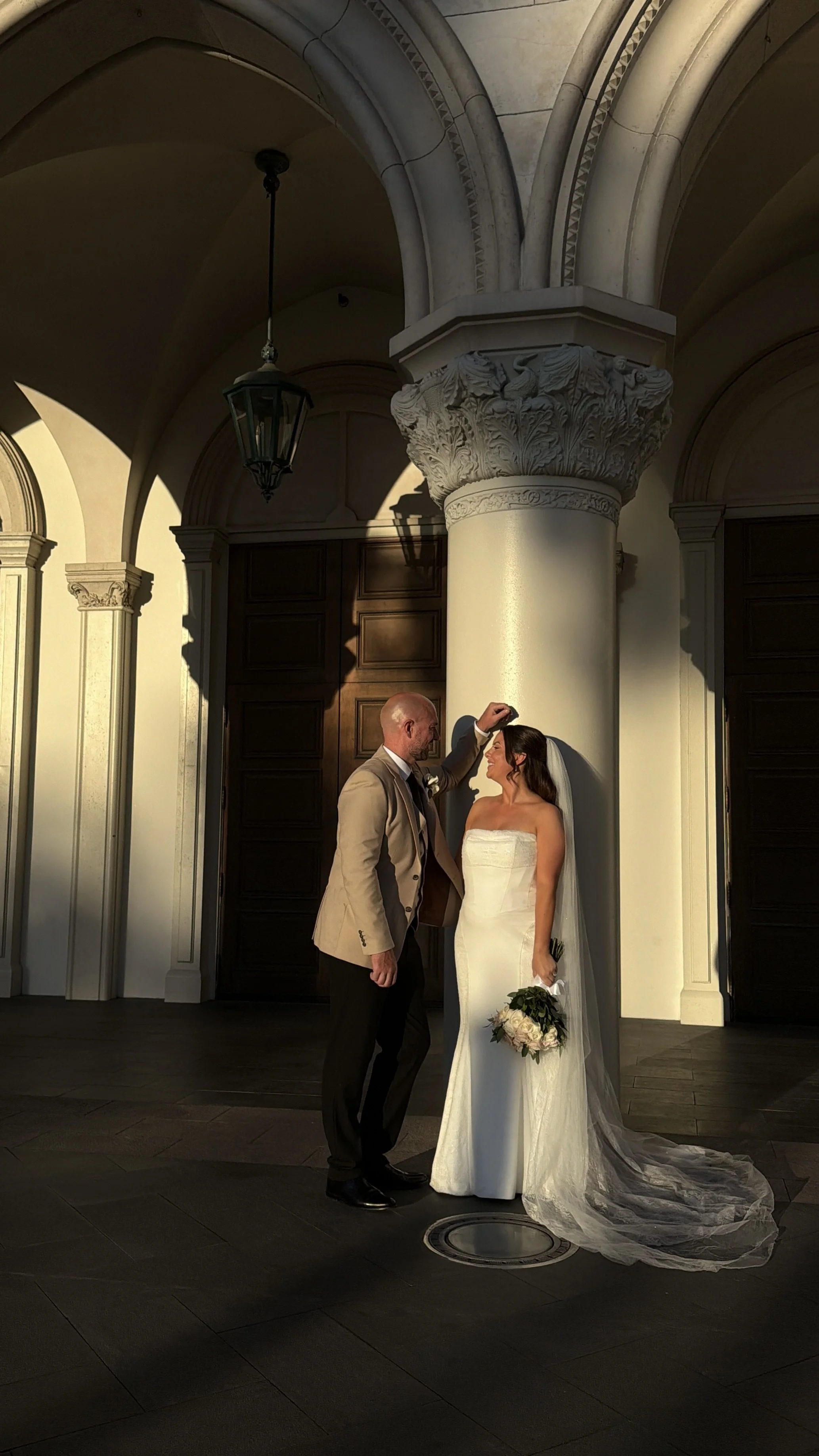 A groom touches a bride's forehead inside a building with classical architecture. The bride is wearing a white wedding dress and holding a bouquet, while the groom is dressed in a beige suit.