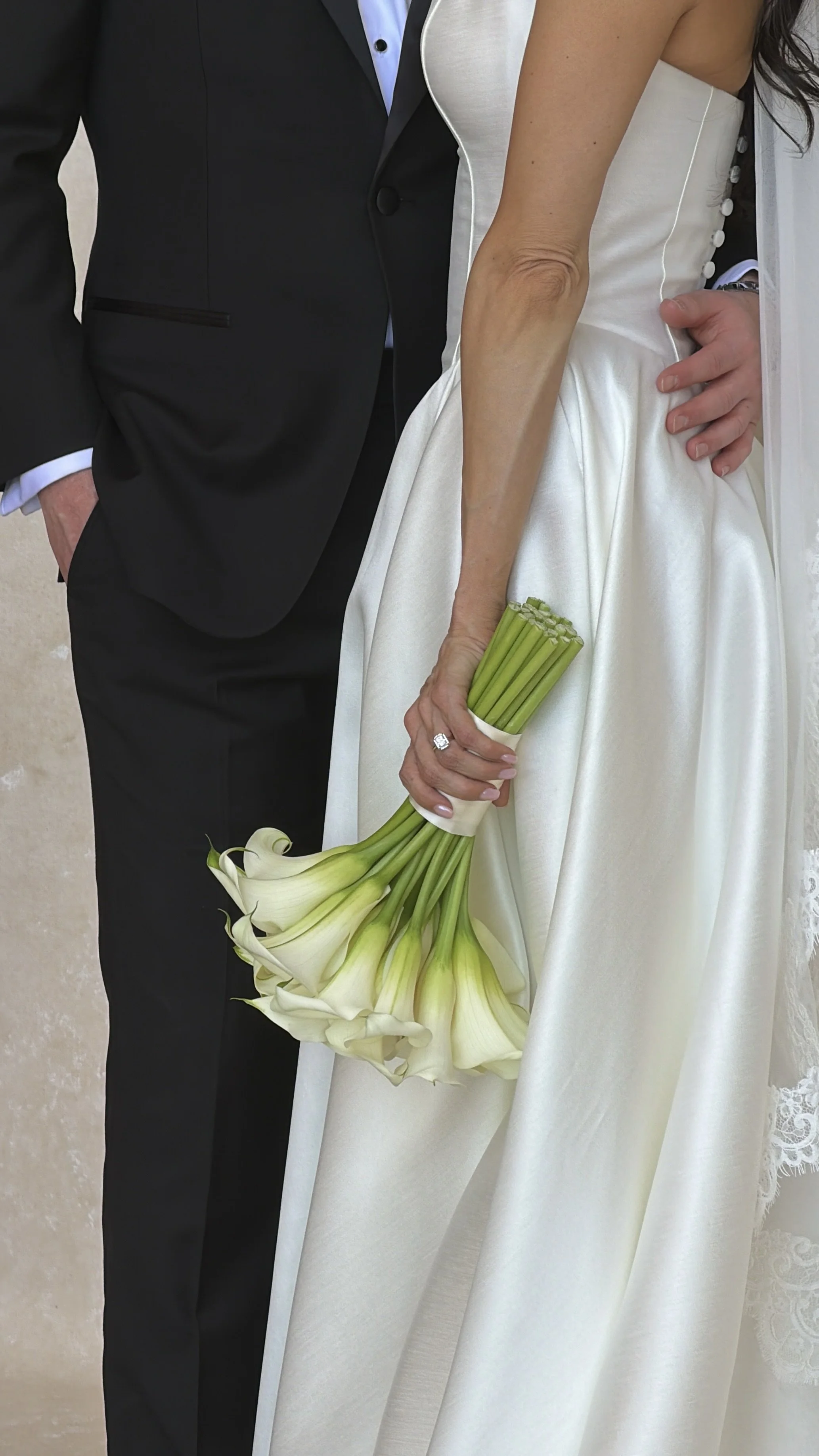 A bride and groom standing close, the bride holding a bouquet of white calla lilies, with the bride in a white wedding dress and the groom in a black tuxedo.