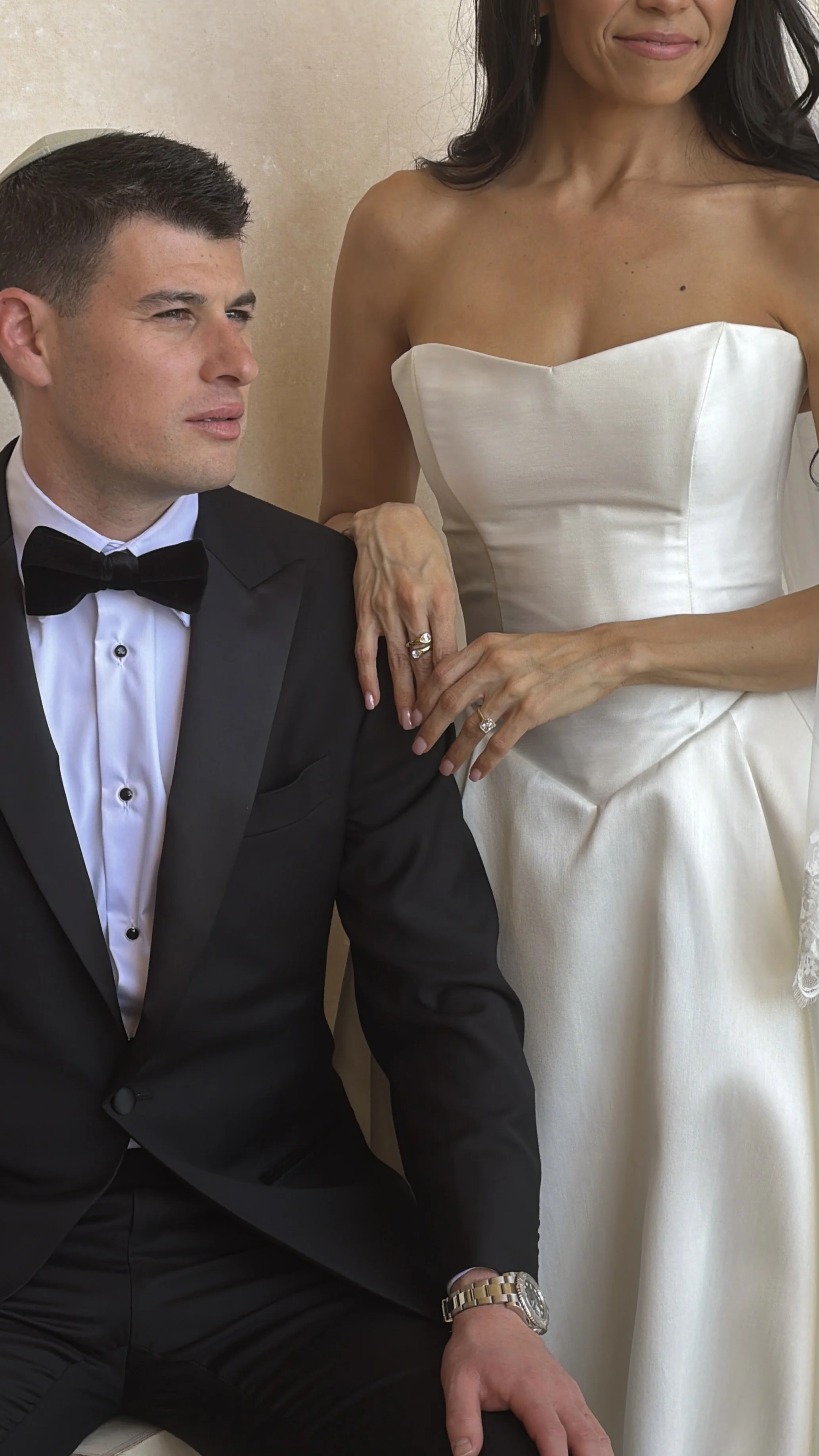 A man in a tuxedo seated next to a woman in a white wedding dress, with her hand resting on his shoulder, showing wedding rings.