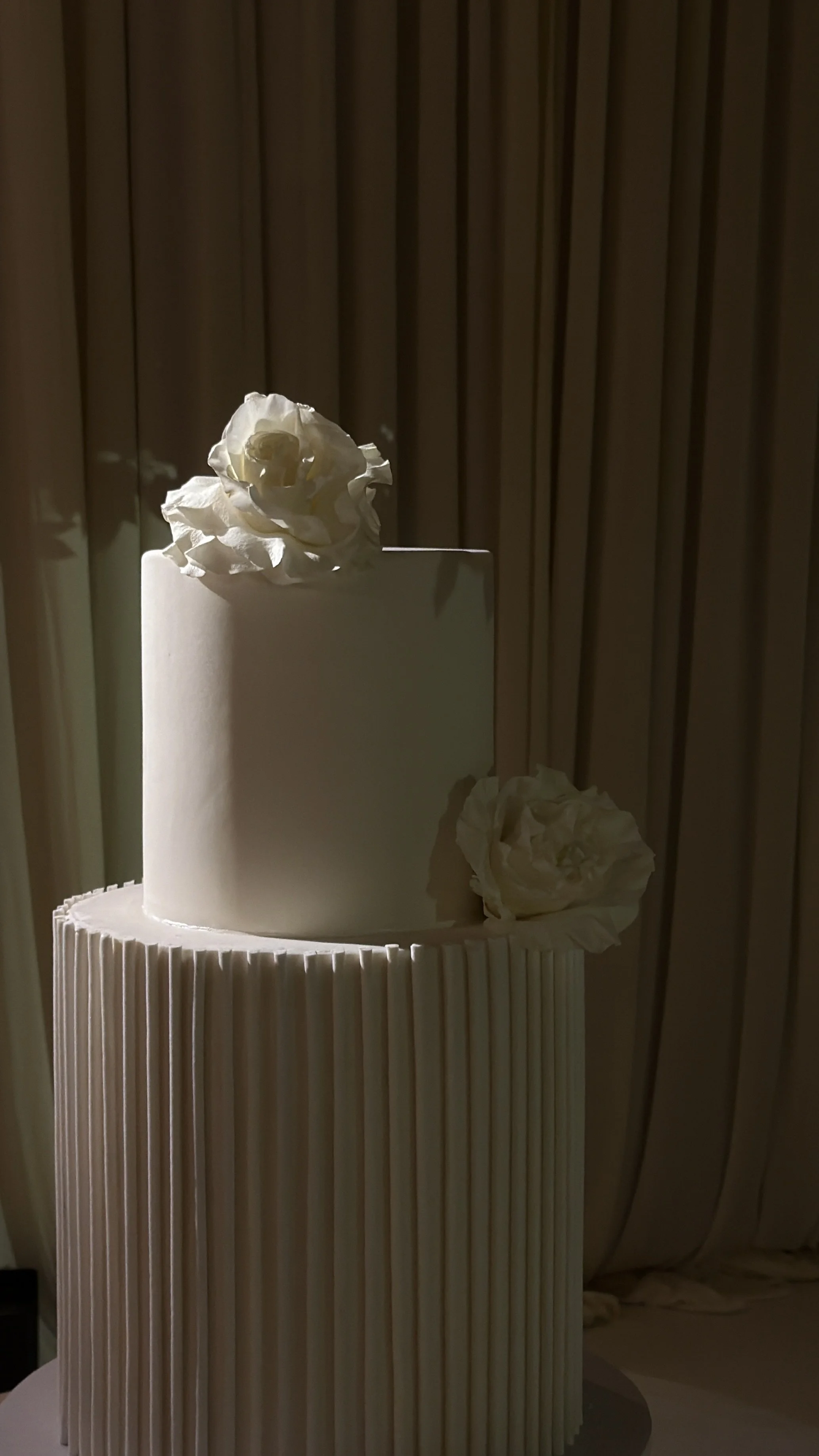 A white tiered cake with vertical grooves and white flowers on top and at the base, with a wooden slat background.