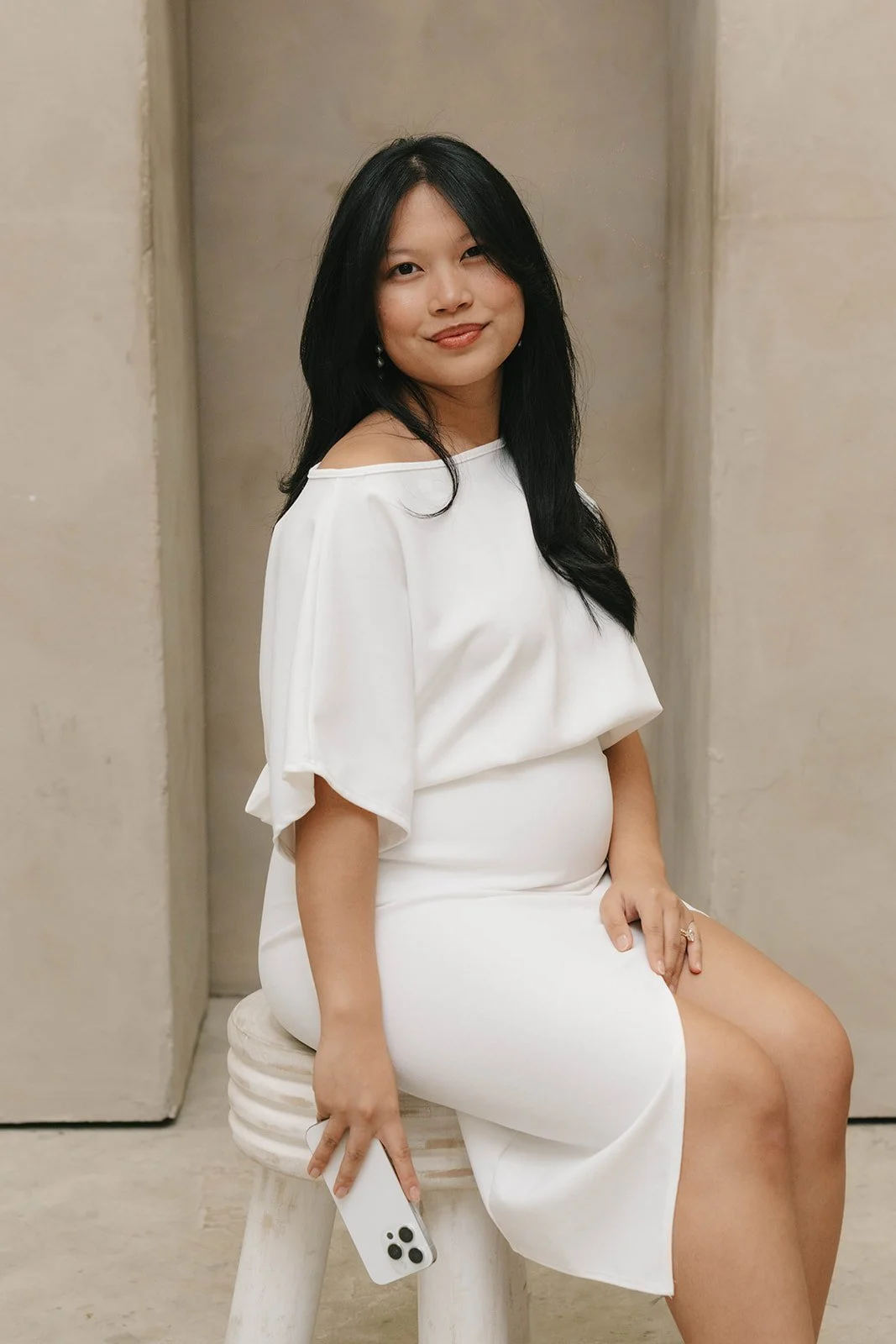 A woman with long black hair is sitting on a white stool in front of a neutral-colored wall. She is wearing a white off-the-shoulder dress, smiling slightly, holding a white smartphone in her left hand, and looking at the camera.