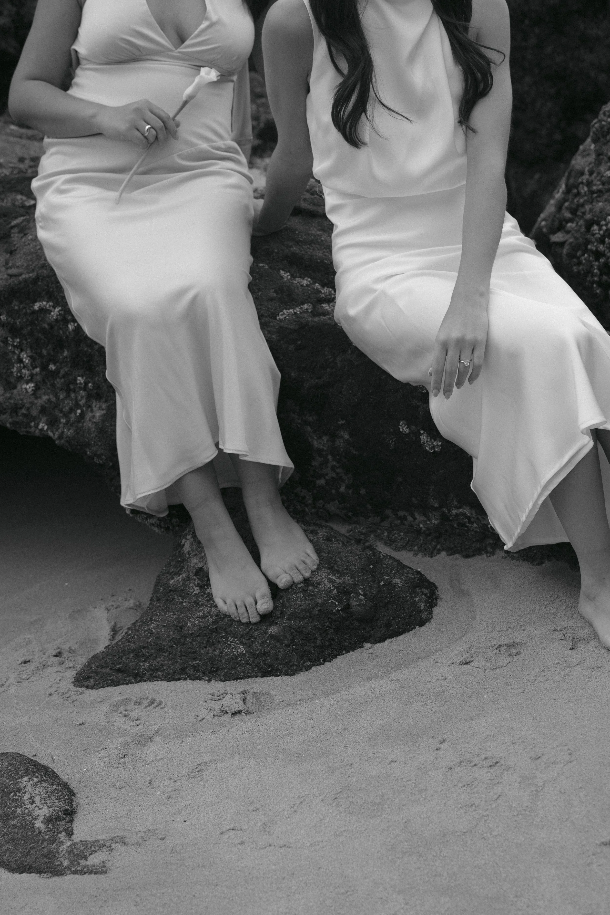 Two women in white dresses sitting on rocks by the beach, showing their legs and feet, with sand and more rocks in the background.