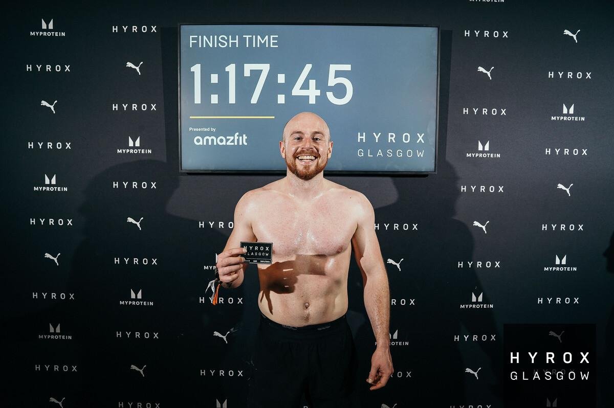 Man with a beard, shirtless, holding a medal, standing in front of a backdrop with logos, after a race, with a digital timer in the background showing 1:17:45.