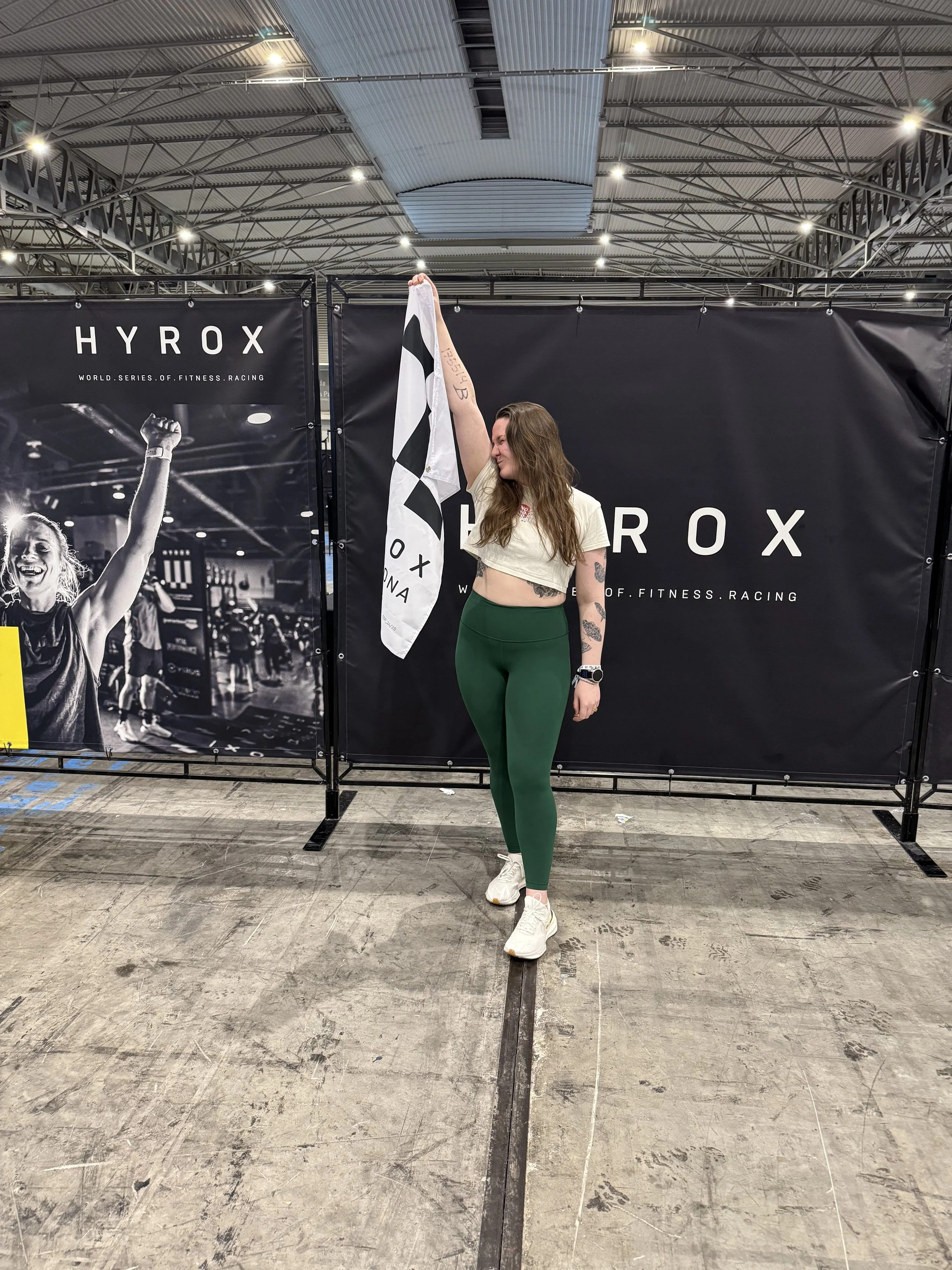 A woman with long brown hair stands in an exhibition hall holding a checkered flag above her head. She is wearing a cropped cream T-shirt, green leggings, and white sneakers. Behind her are black banners with the text 'HYROX' and a black and white photograph of a woman celebrating.