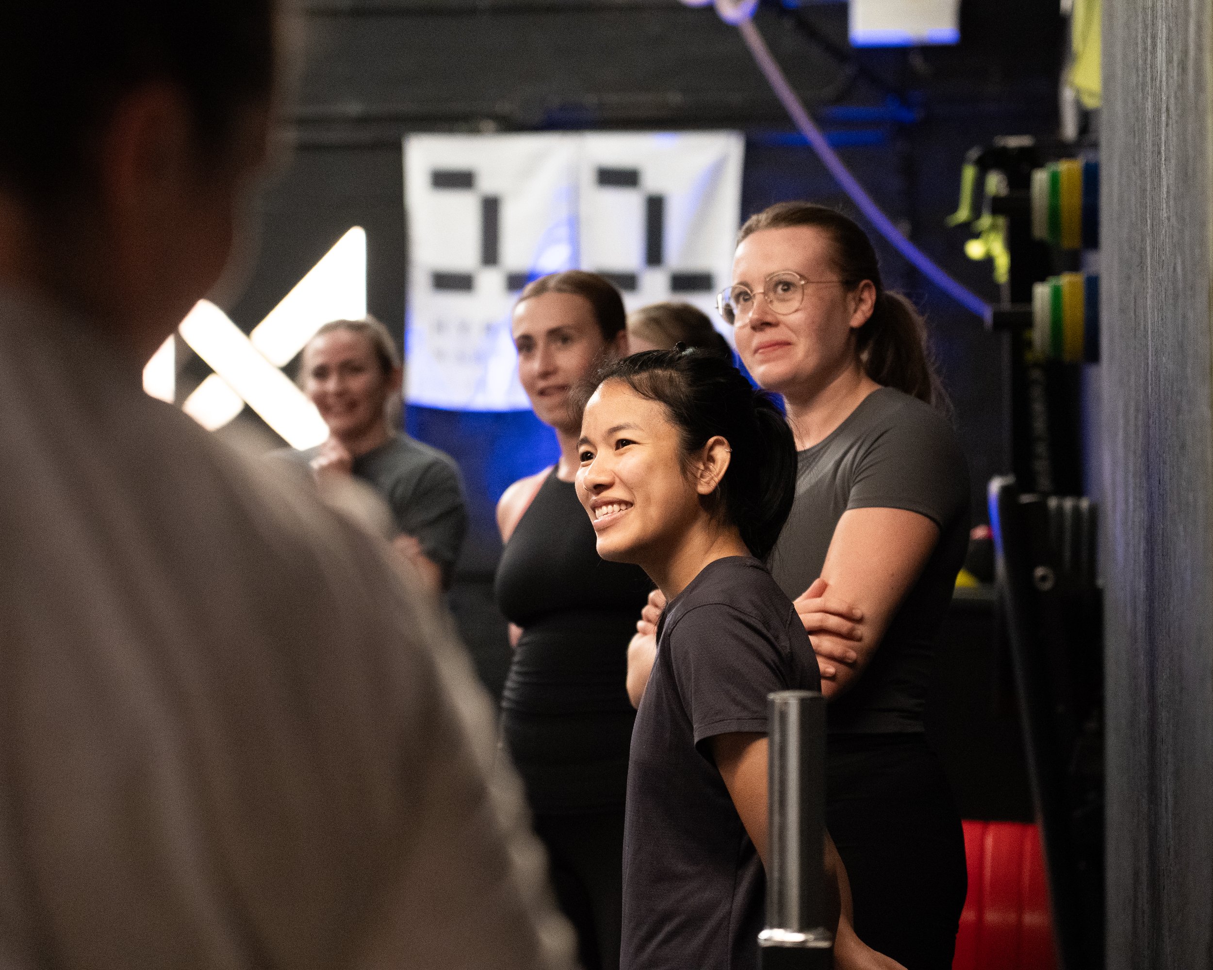 A group of women, including one Asian woman smiling, are standing together in what appears to be a gym or fitness studio, with exercise equipment visible in the background.