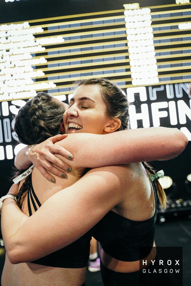 Two women hugging and smiling in a gym, with fitness tracking boards in the background, at Hyrox Glasgow event.