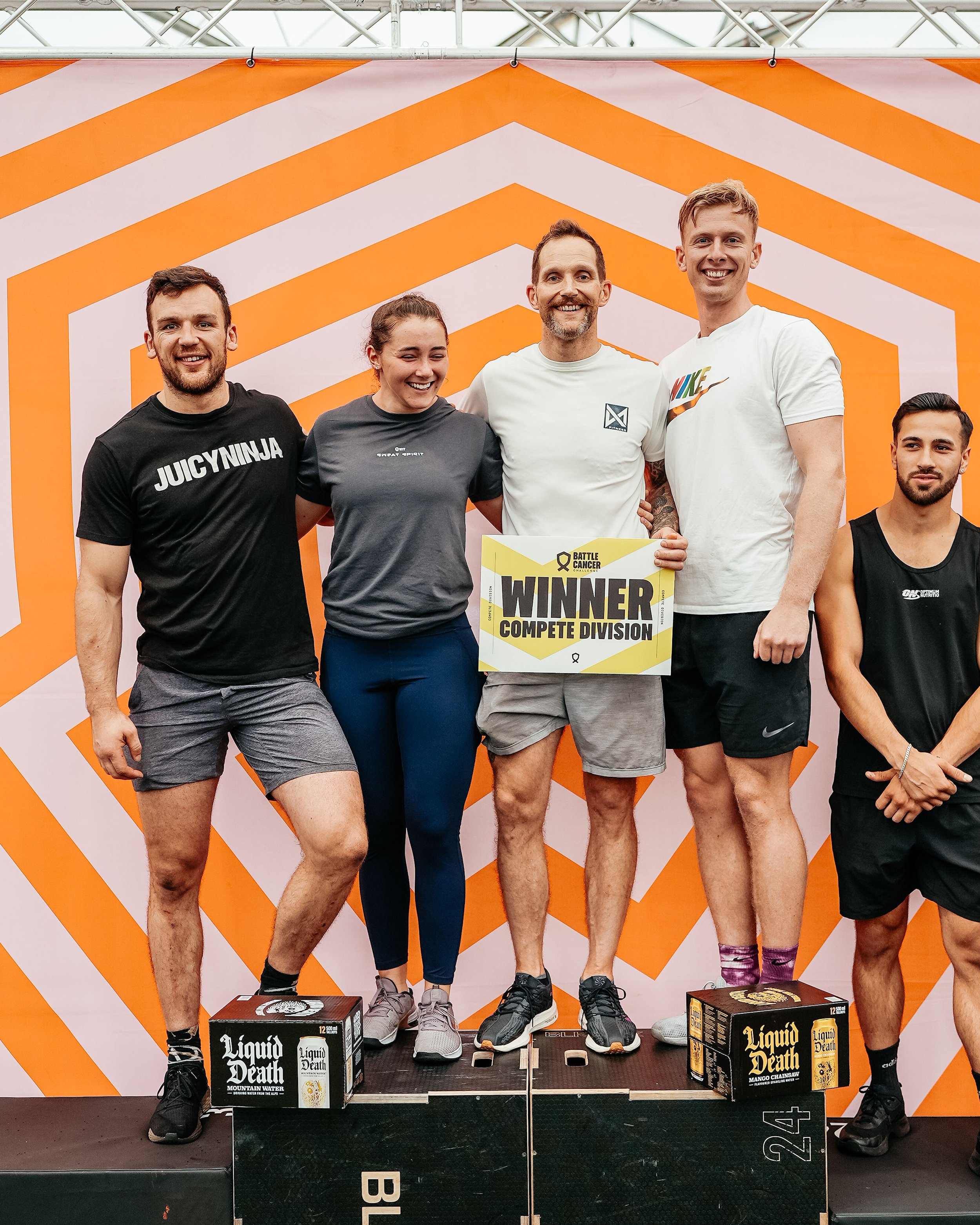 Group of five people on a podium celebrating a victory at a fitness event, holding a sign that says 'Winner, Compete Division,' with a backdrop featuring orange and white geometric patterns.