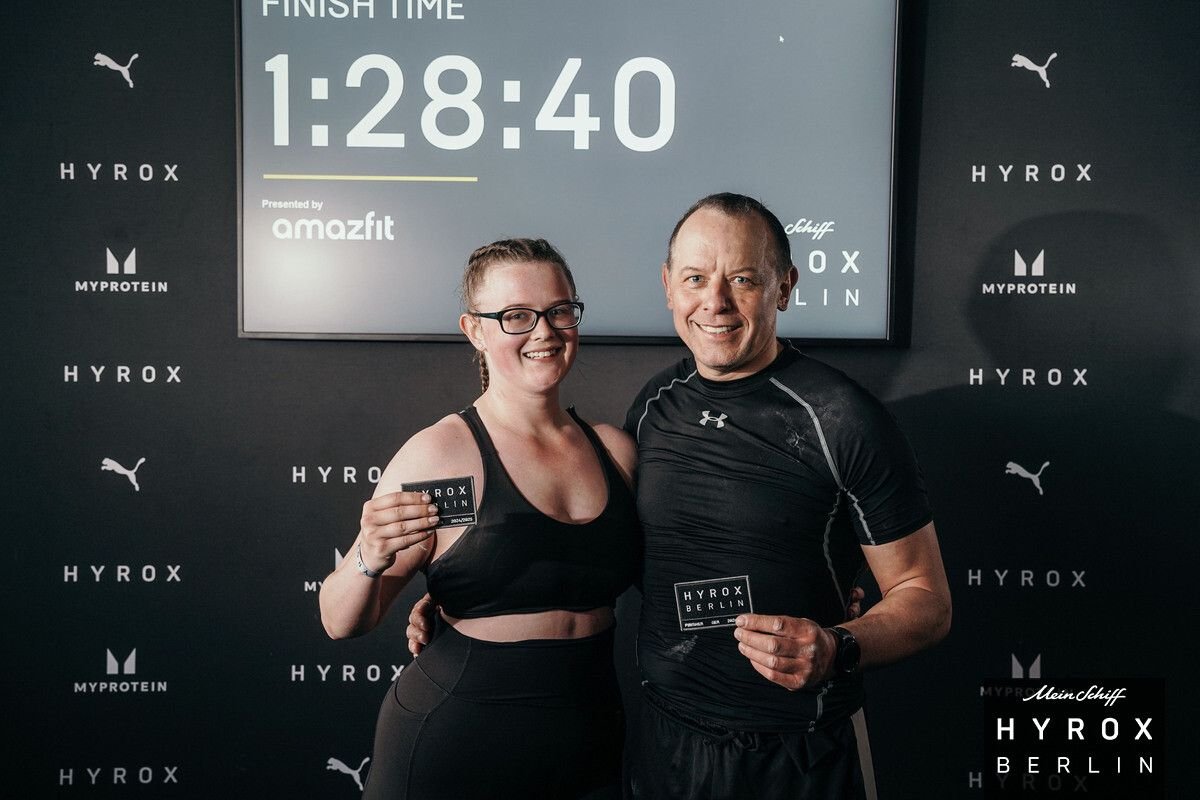 Two people, a woman and a man, smiling and holding medals, standing in front of a fitness event backdrop with a large digital timer showing 1:28:40, and logos of Hyrox, Puma, and MyProtein.
