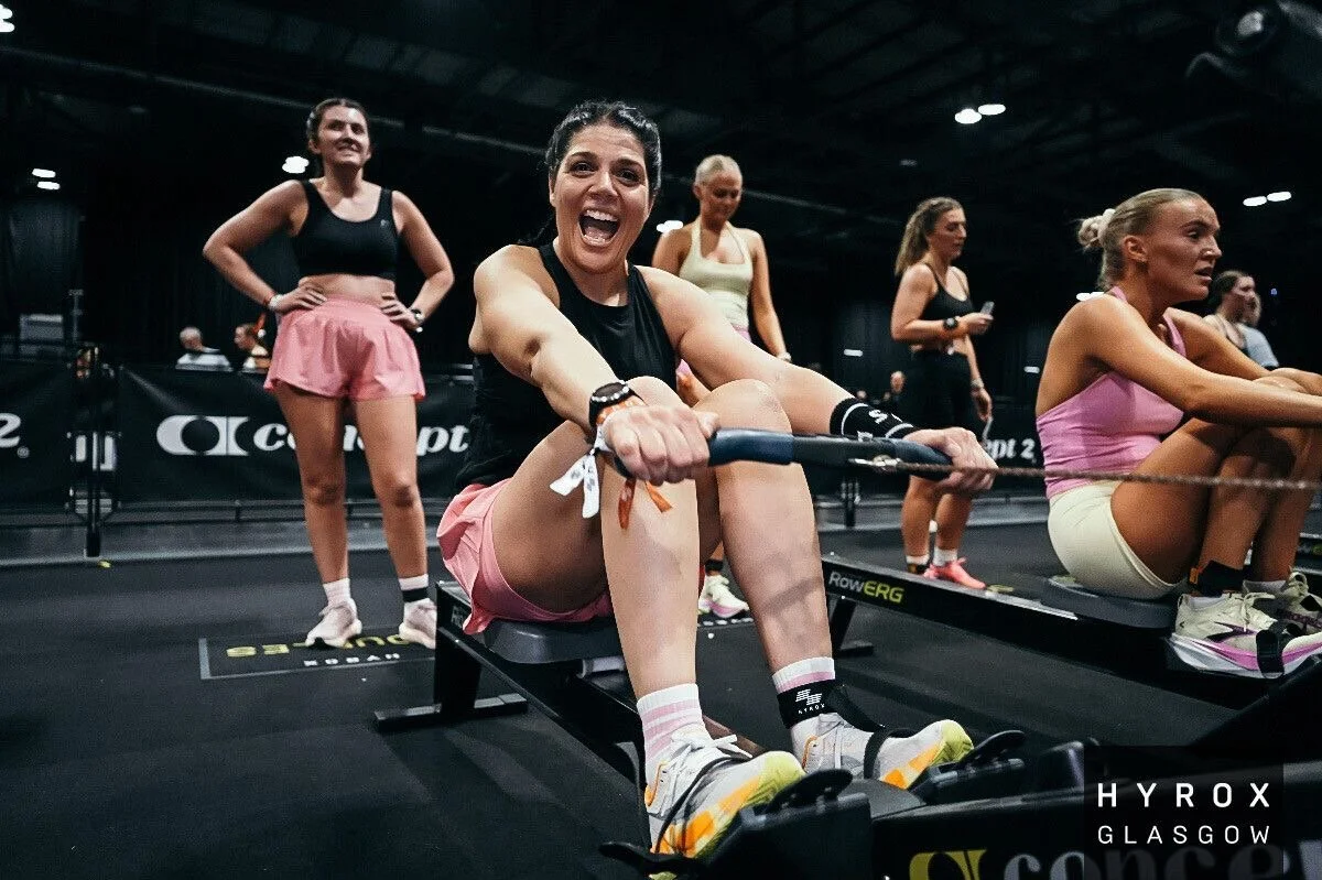 A woman running on a trail in a wooded area, smiling and waving, wearing a black tank top, patterned leggings, and a bib numbered 193.