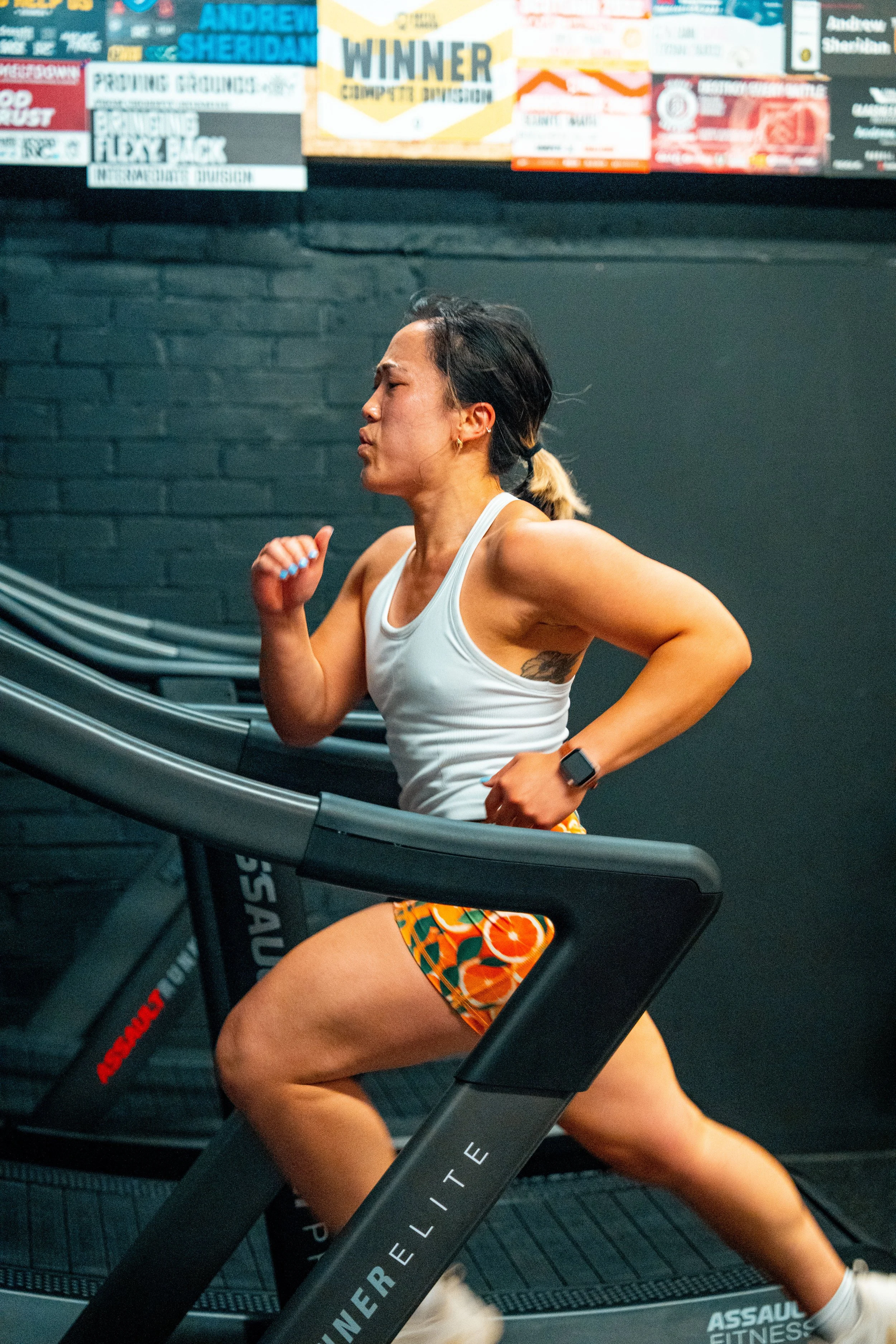 A woman running on a treadmill in a gym, wearing a white tank top and colorful shorts with orange and green patterns, with a black fitness watch on her left wrist, and a black hair tie in her hair, behind a black wall with various posters and signs.